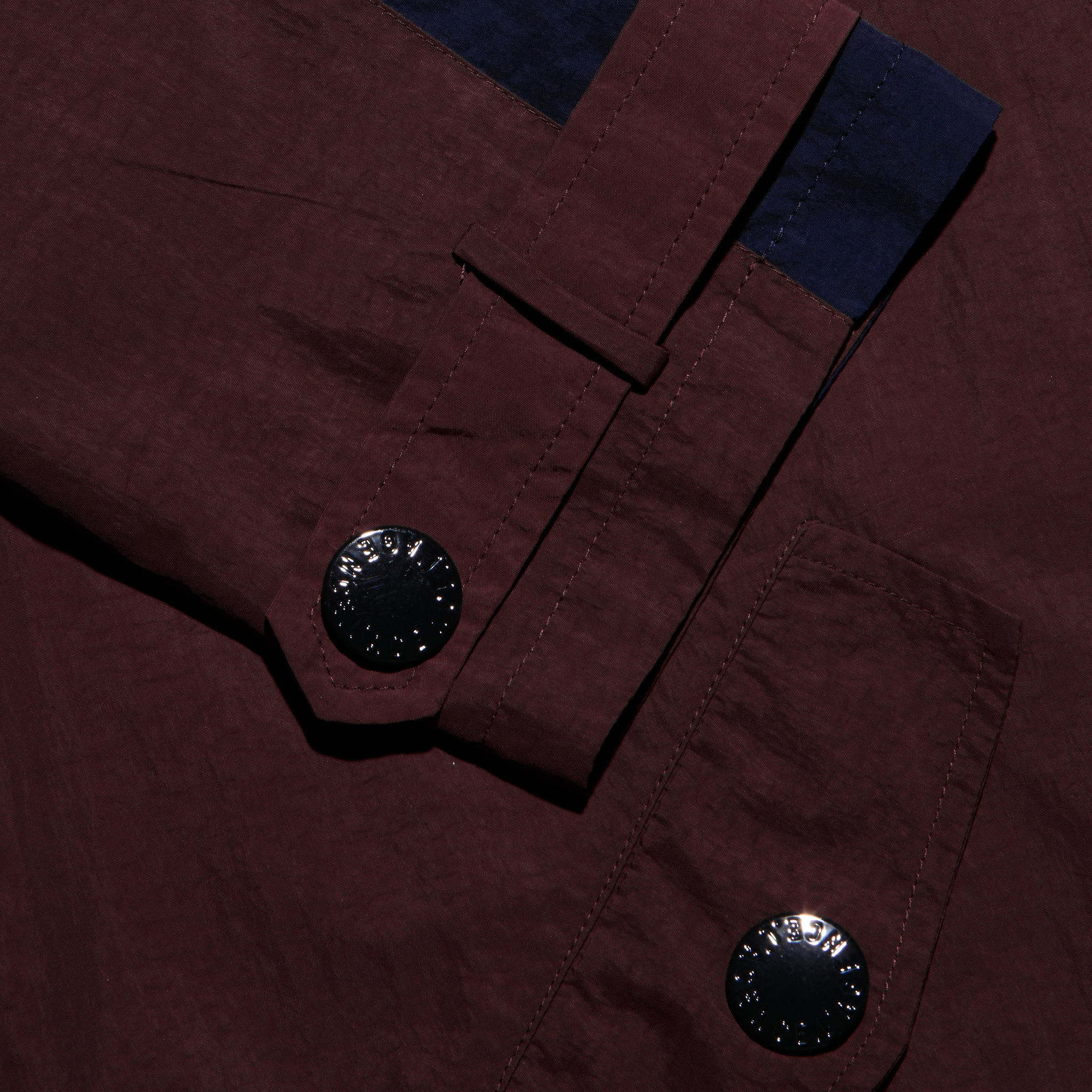 Maroon Geovana windbreaker sleeve featuring a dark blue cuff accent, a strap with a black button, and a large black button on the water-repellant pocket.