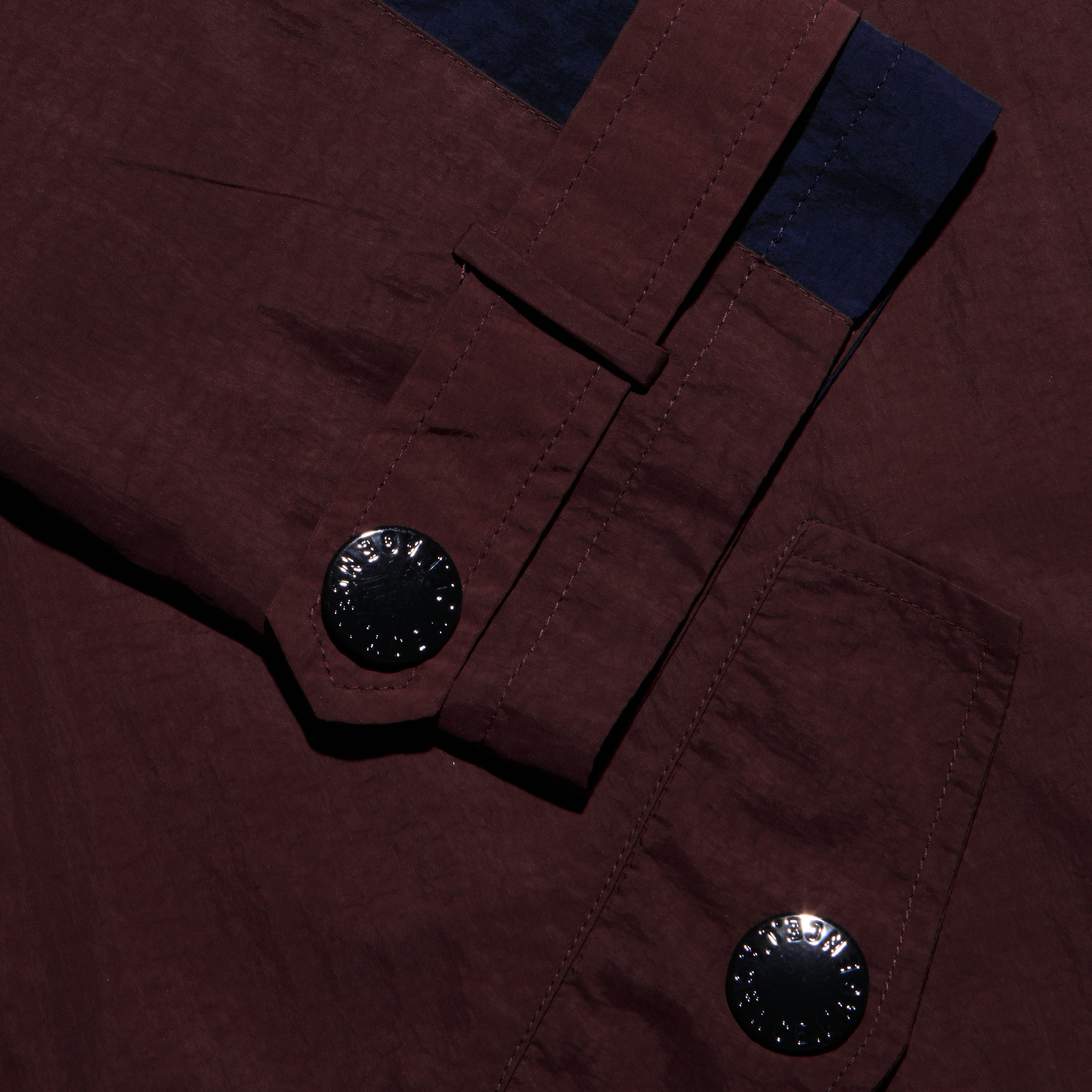 Maroon Geovana windbreaker sleeve featuring a dark blue cuff accent, a strap with a black button, and a large black button on the water-repellant pocket.