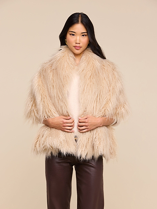 A woman stands facing the camera, wearing the Sabine Faux Fur Scarf—an elegant cool-weather accessory—in beige draped over a light top and dark brown pants, with her hands gently holding the scarf against a plain beige background.