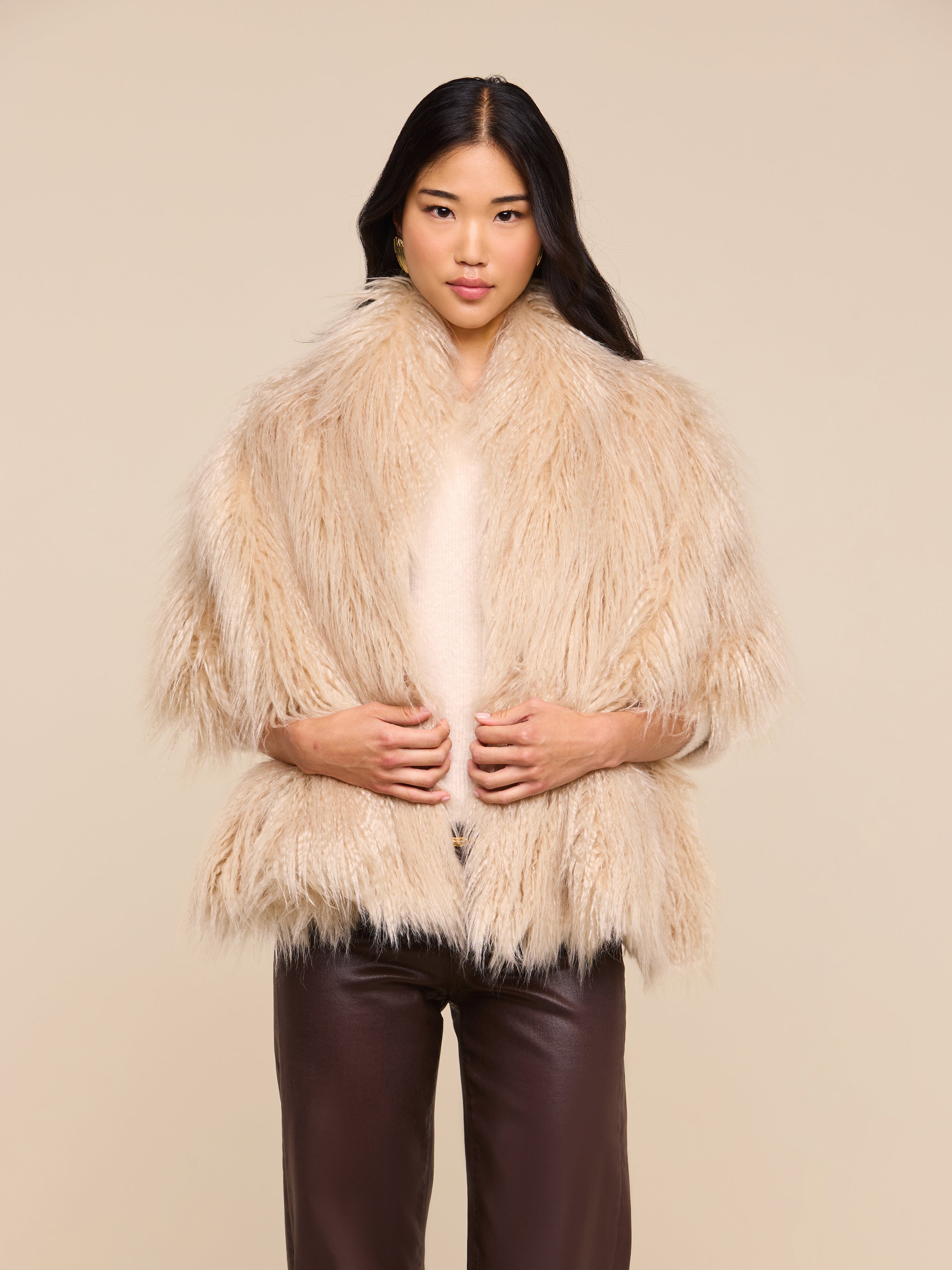 A woman stands facing the camera, wearing the Sabine Faux Fur Scarf—an elegant cool-weather accessory—in beige draped over a light top and dark brown pants, with her hands gently holding the scarf against a plain beige background.