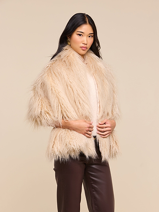 A woman with long black hair wears the Sabine Faux Fur Scarf—a perfect cool-weather accessory—over a light top and brown leather pants, standing against a plain beige background.