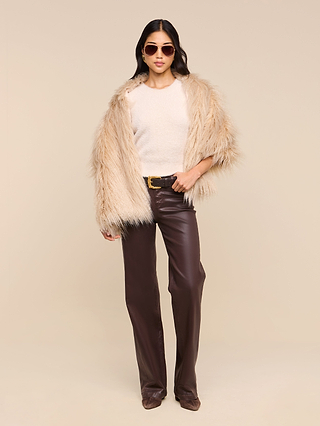 A woman wearing sunglasses drapes the Sabine Faux Fur Scarf stylishly over her shoulders as a chic cool-weather accessory, paired with a cream sweater and high-waisted brown leather pants, standing confidently against a plain beige background.