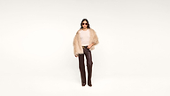 A person wearing sunglasses, a beige fur coat, a white top, and brown leather pants stands confidently against a plain white background.