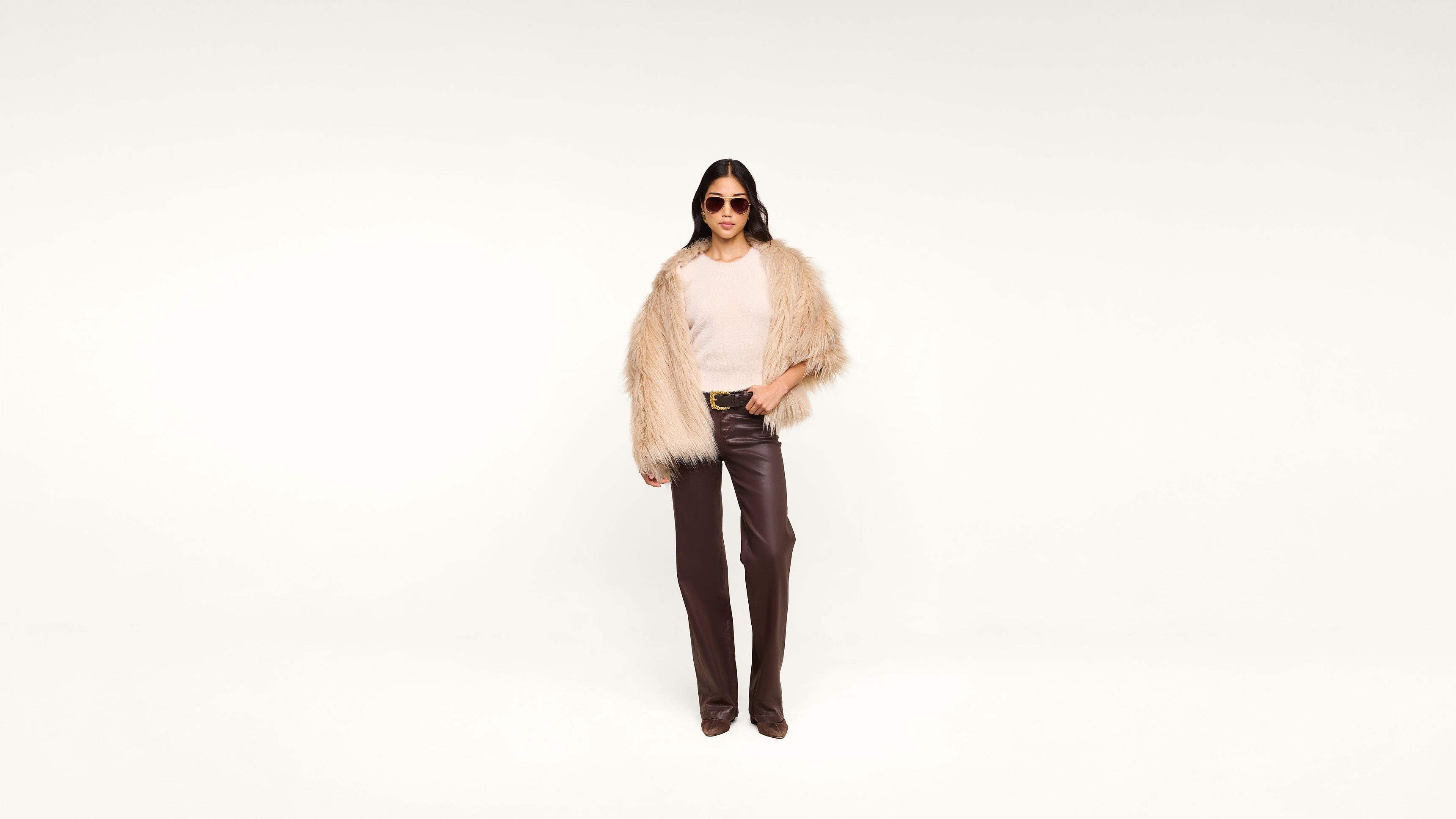 A person wearing sunglasses, a beige fur coat, a white top, and brown leather pants stands confidently against a plain white background.