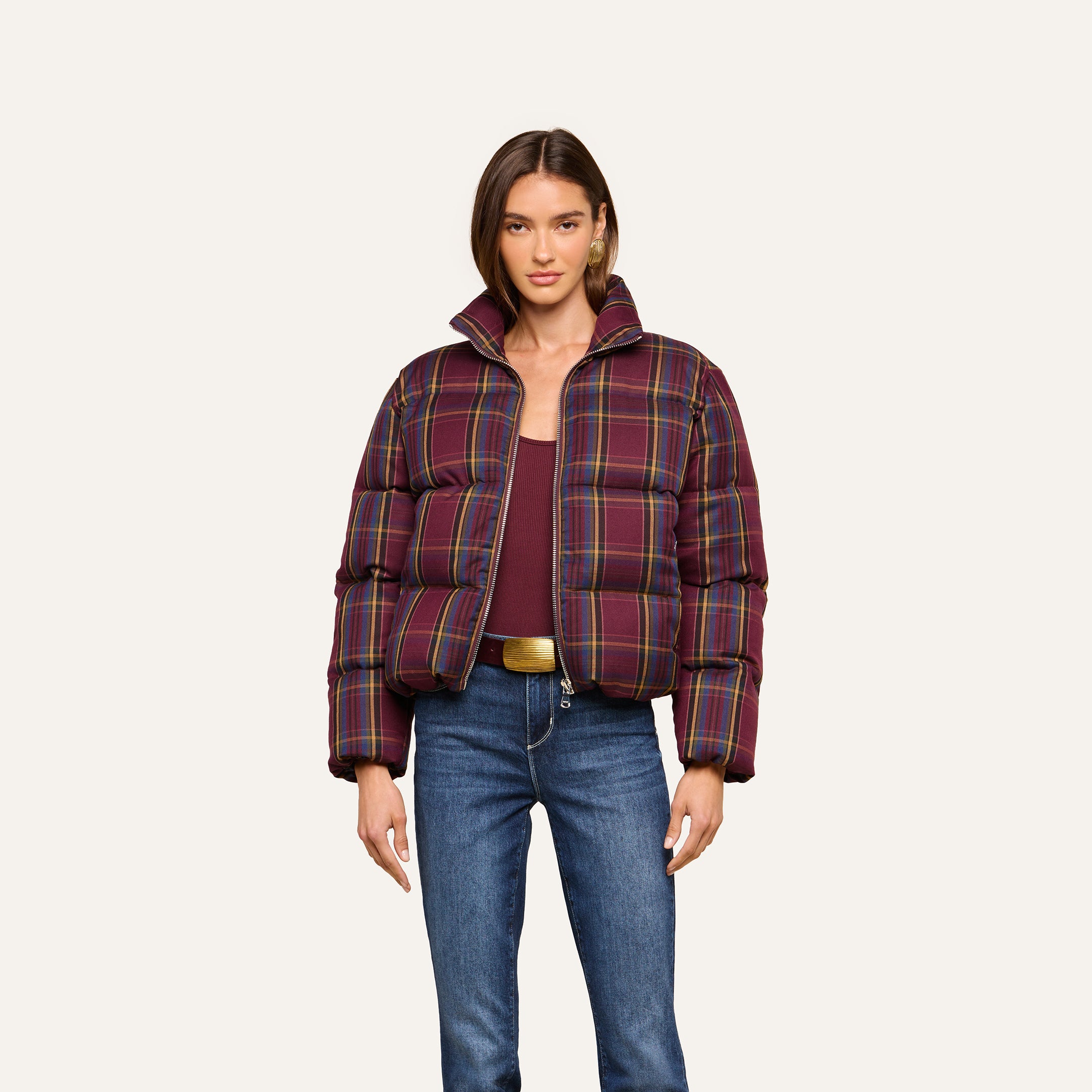 A woman wearing the Larsa Oversized Puffer Jacket over a maroon top, blue jeans, and a beige belt, standing against a plain light background.