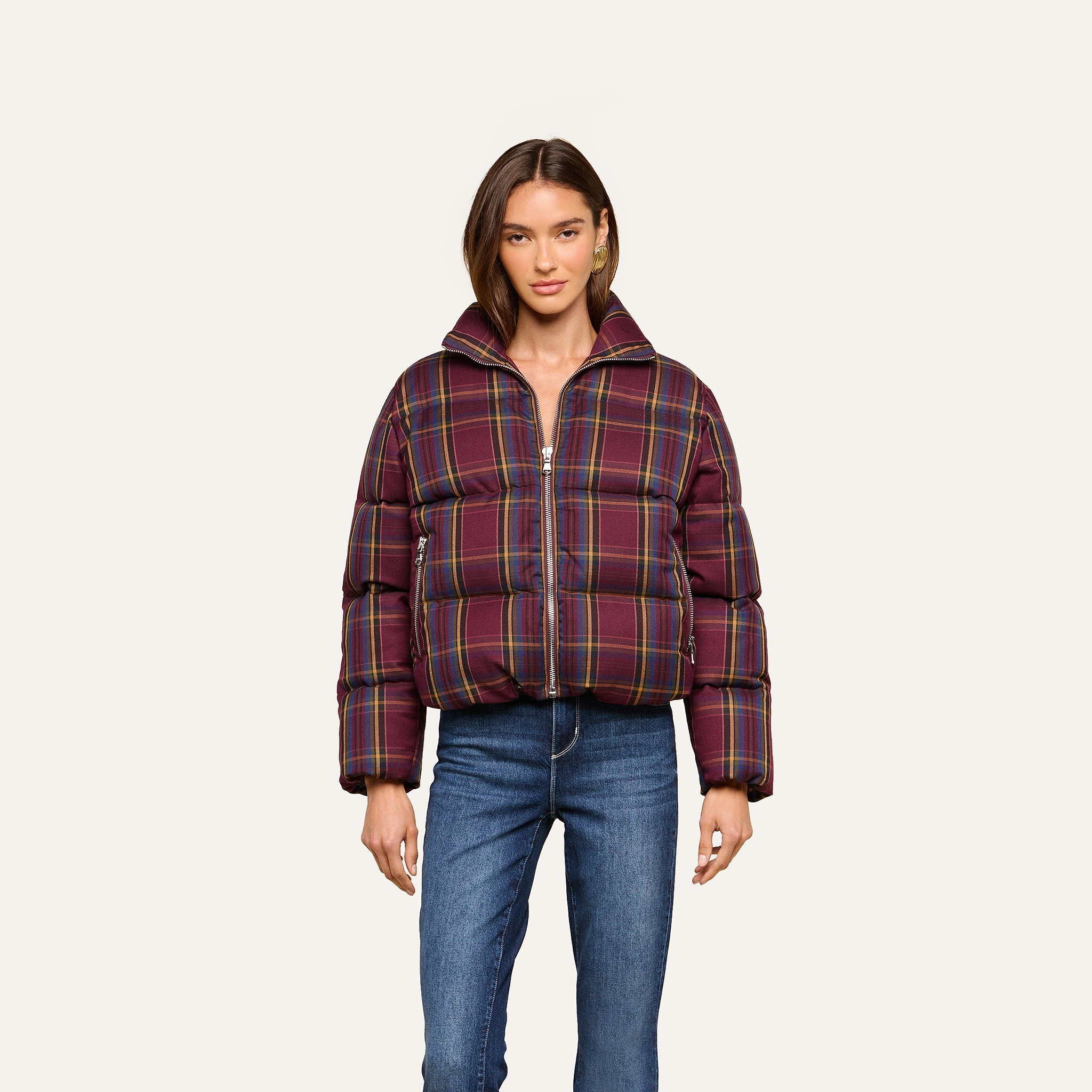 A woman stands against a plain light background, facing forward with her arms relaxed by her sides, wearing the Larsa Oversized Puffer Jacket in purple plaid with a high collar, paired with blue jeans.