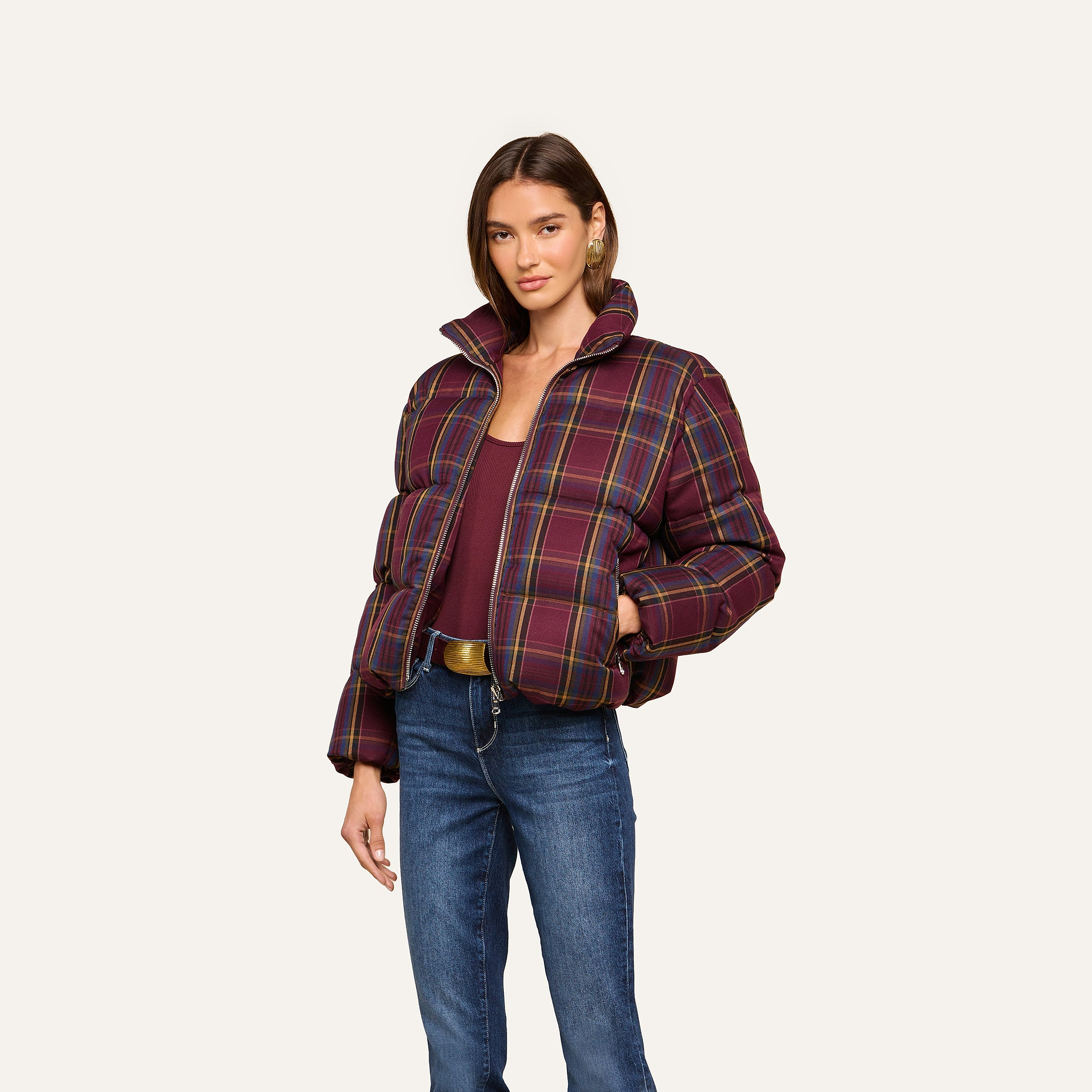 A woman wearing the Larsa Oversized Puffer Jacket, a maroon top, blue jeans, and a brown belt stands against a plain light background.