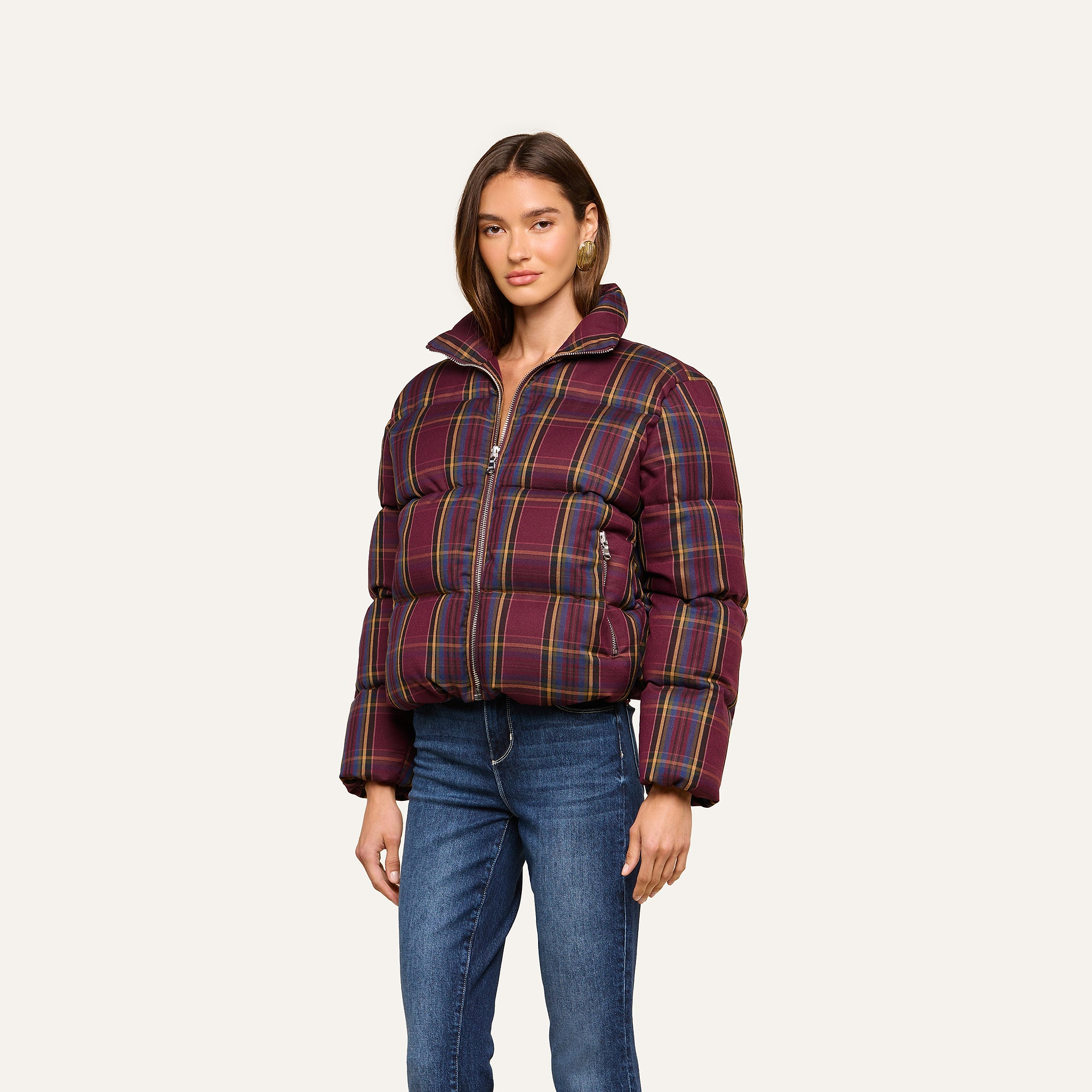 A woman wearing the Larsa Oversized Puffer Jacket in maroon plaid, paired with blue jeans and gold hoop earrings, stands against a plain light background.