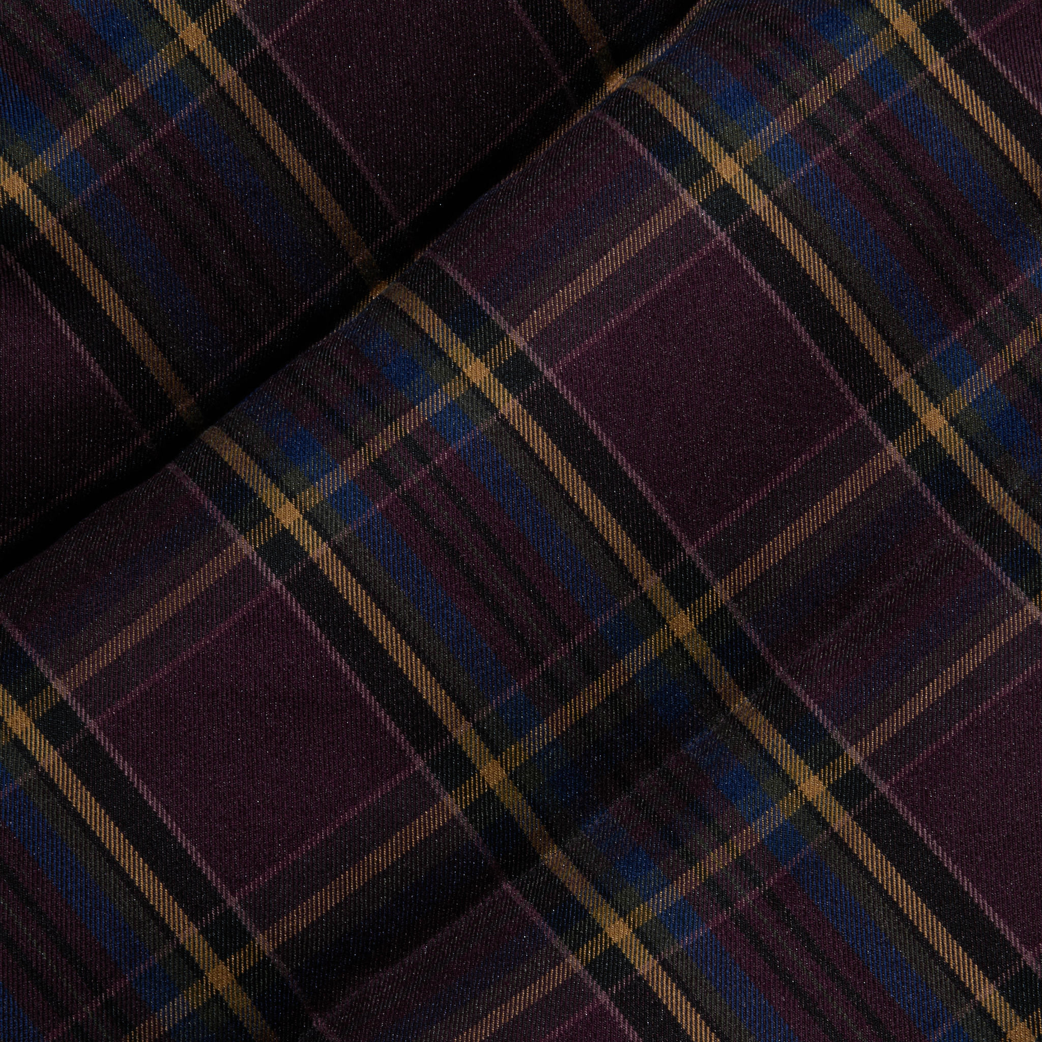 The Larsa Oversized Puffer Jacket features dark purple tartan fabric with intersecting lines in gold, black, and blue, creating a classic plaid pattern. The soft woven texture recalls traditional plaid materials, with two sections of the fabric slightly overlapping at an angle.