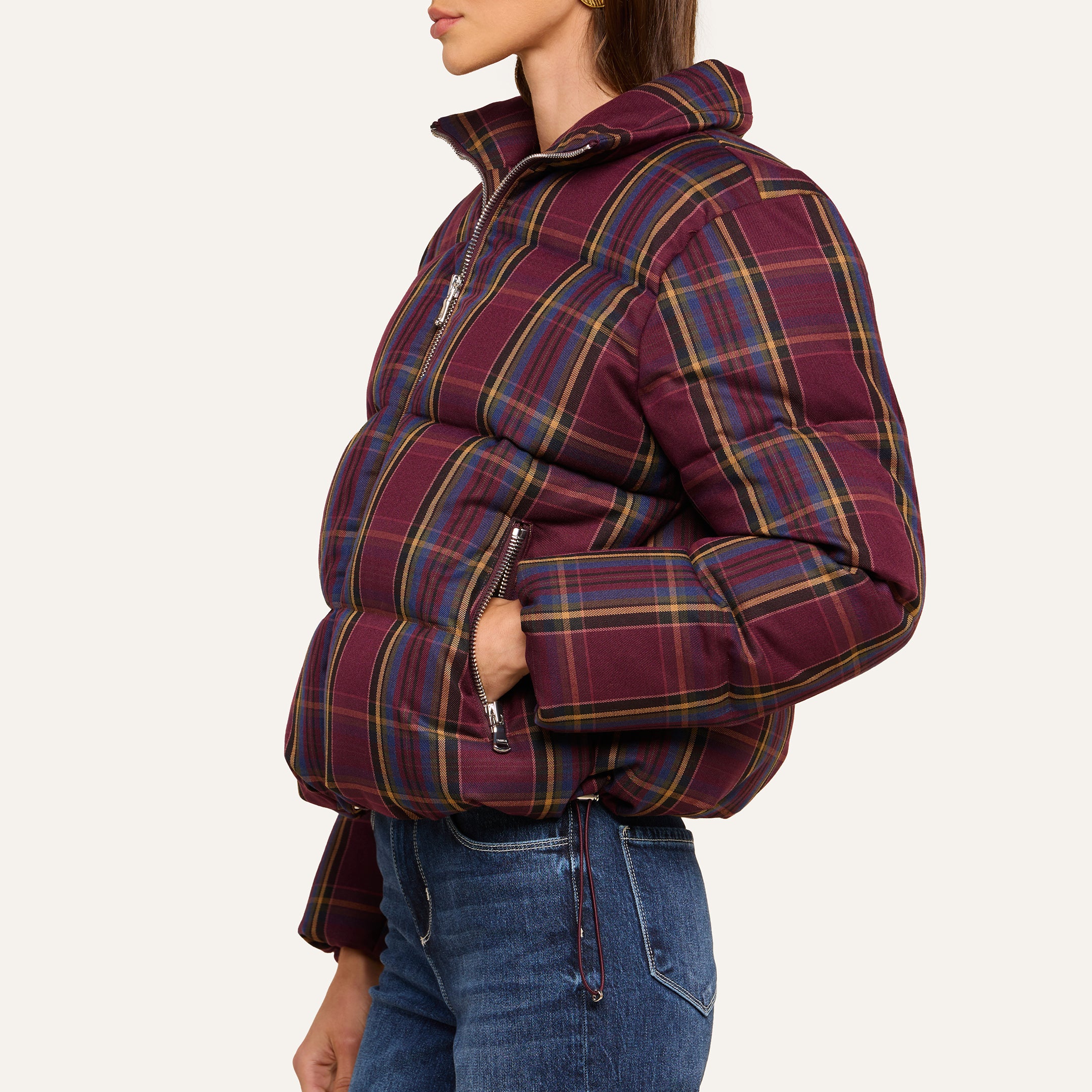 A woman stands in profile against a plain background wearing the Larsa Oversized Puffer Jacket in purple plaid with zipper pockets, paired with blue jeans.