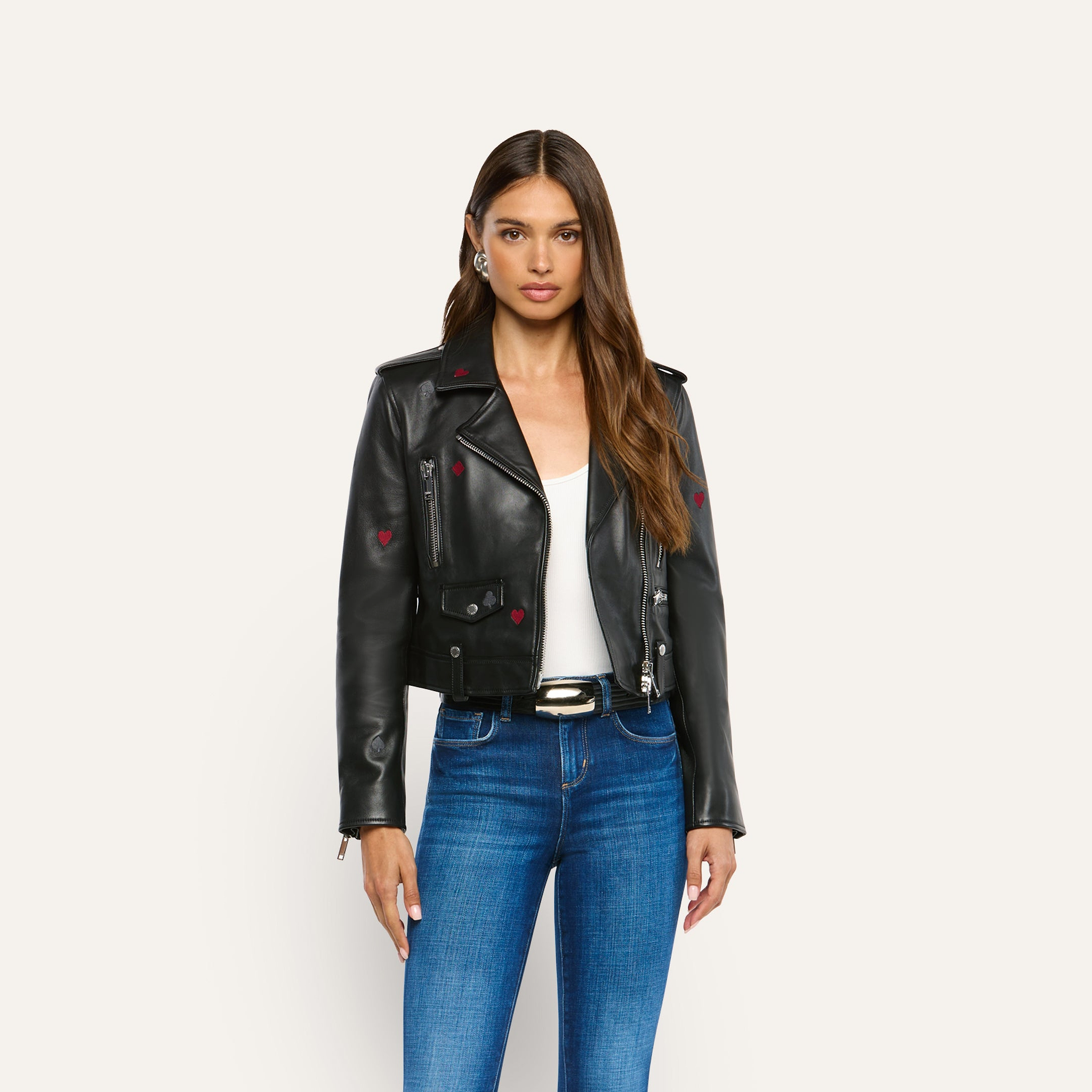 A woman wears a Blakely Embroidered Leather Jacket in black lambskin with small red heart details over a white top and blue jeans.