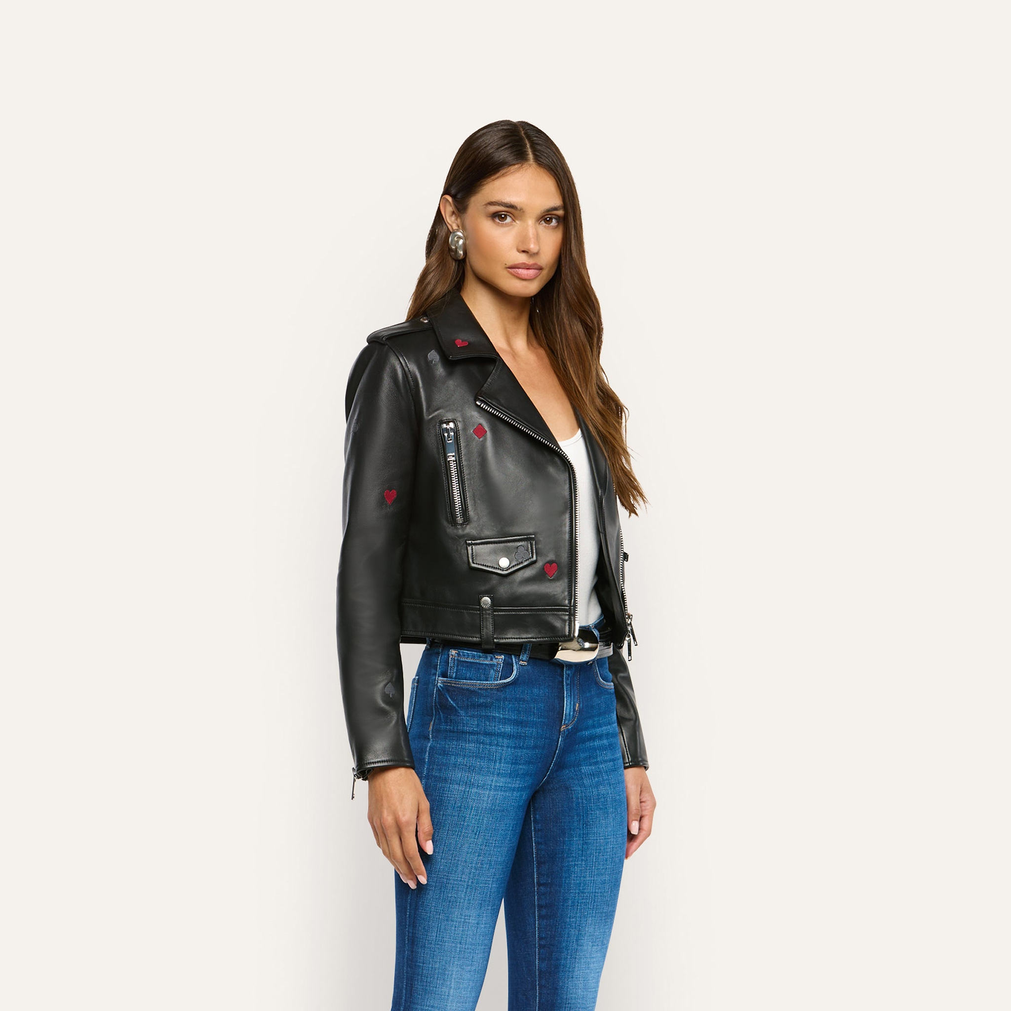 A woman wears a black lambskin Blakely Embroidered Leather Jacket featuring small red embroidered heart accents, paired with a white top and blue jeans.