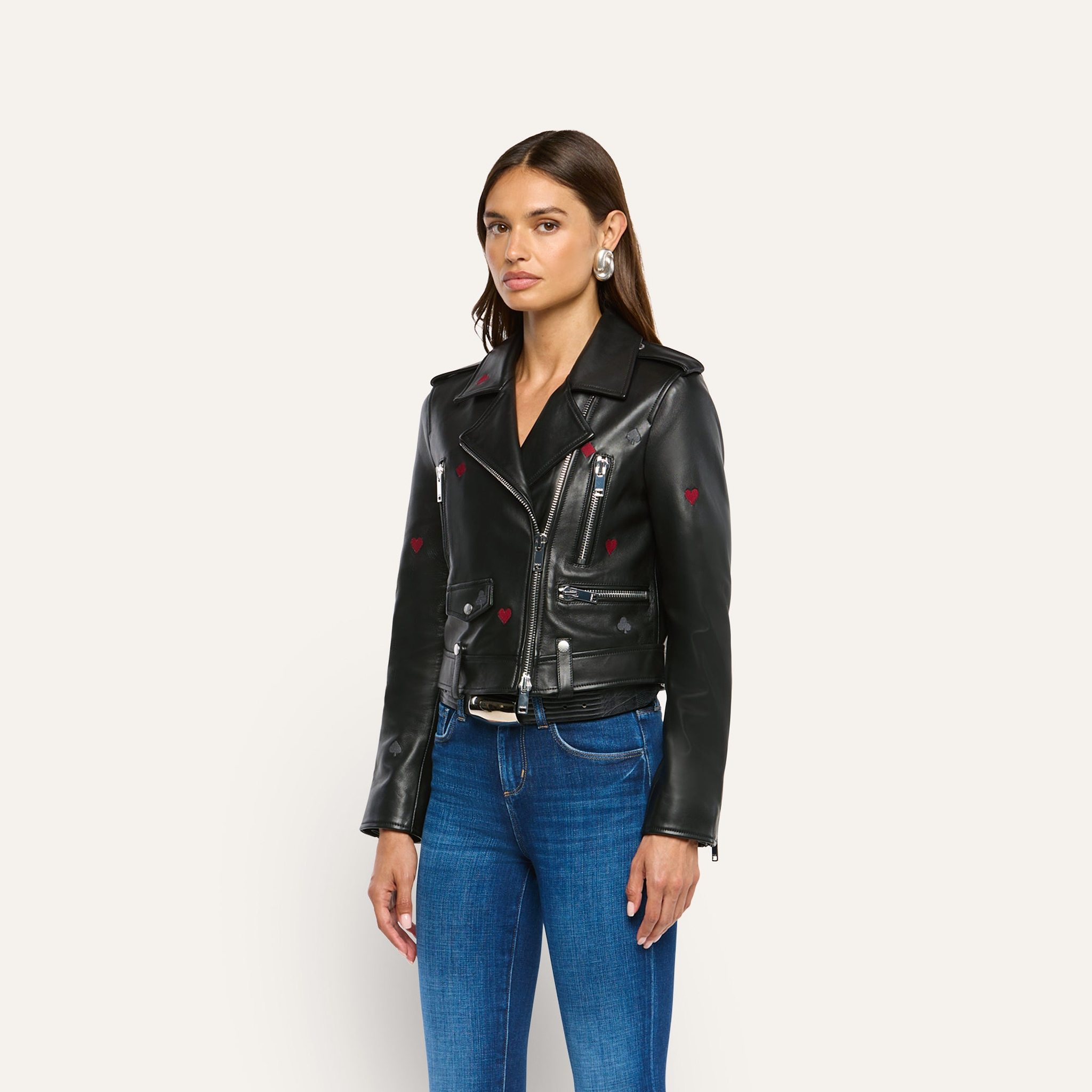A model wears a Blakely Embroidered Leather Jacket, an embroidered lambskin biker jacket featuring small red hearts and black spade symbols, paired with blue jeans.