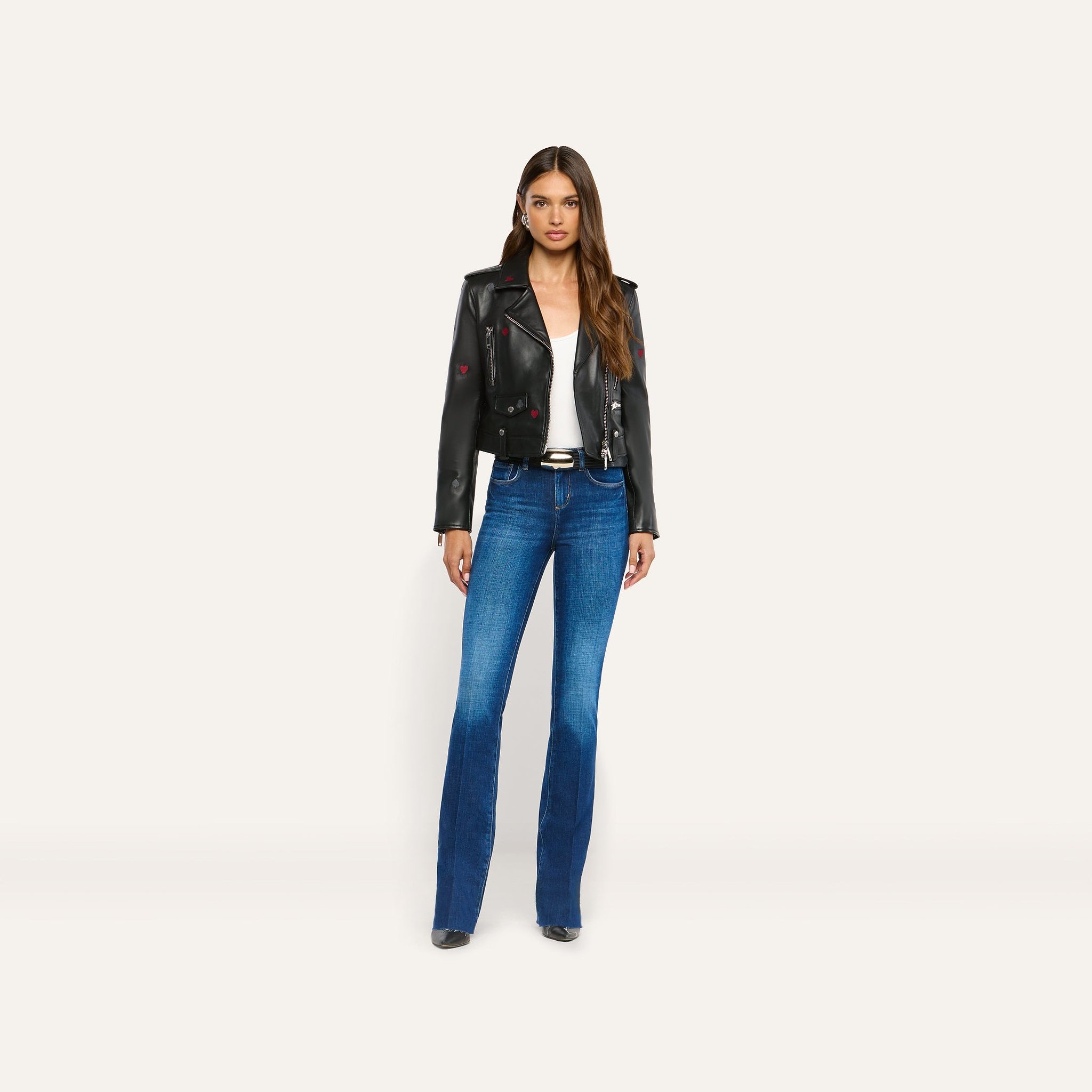 A woman wears a black lambskin Blakely Embroidered Leather Jacket featuring small red embroidered hearts, a white tank top, and blue flared jeans.