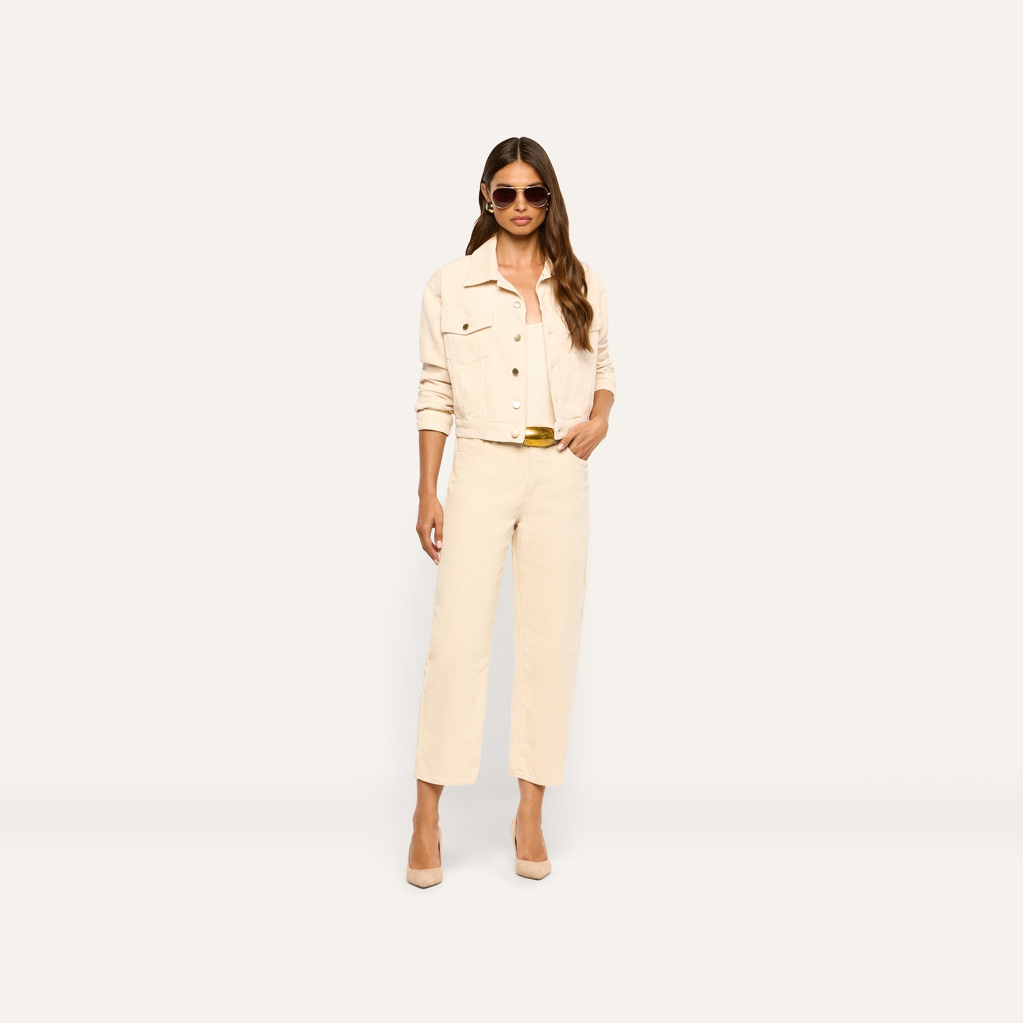 A woman wears an Alessia Corduroy Jacket in pearl white with matching high-waisted pants, a gold-buckled belt, tan pumps, and sunglasses.