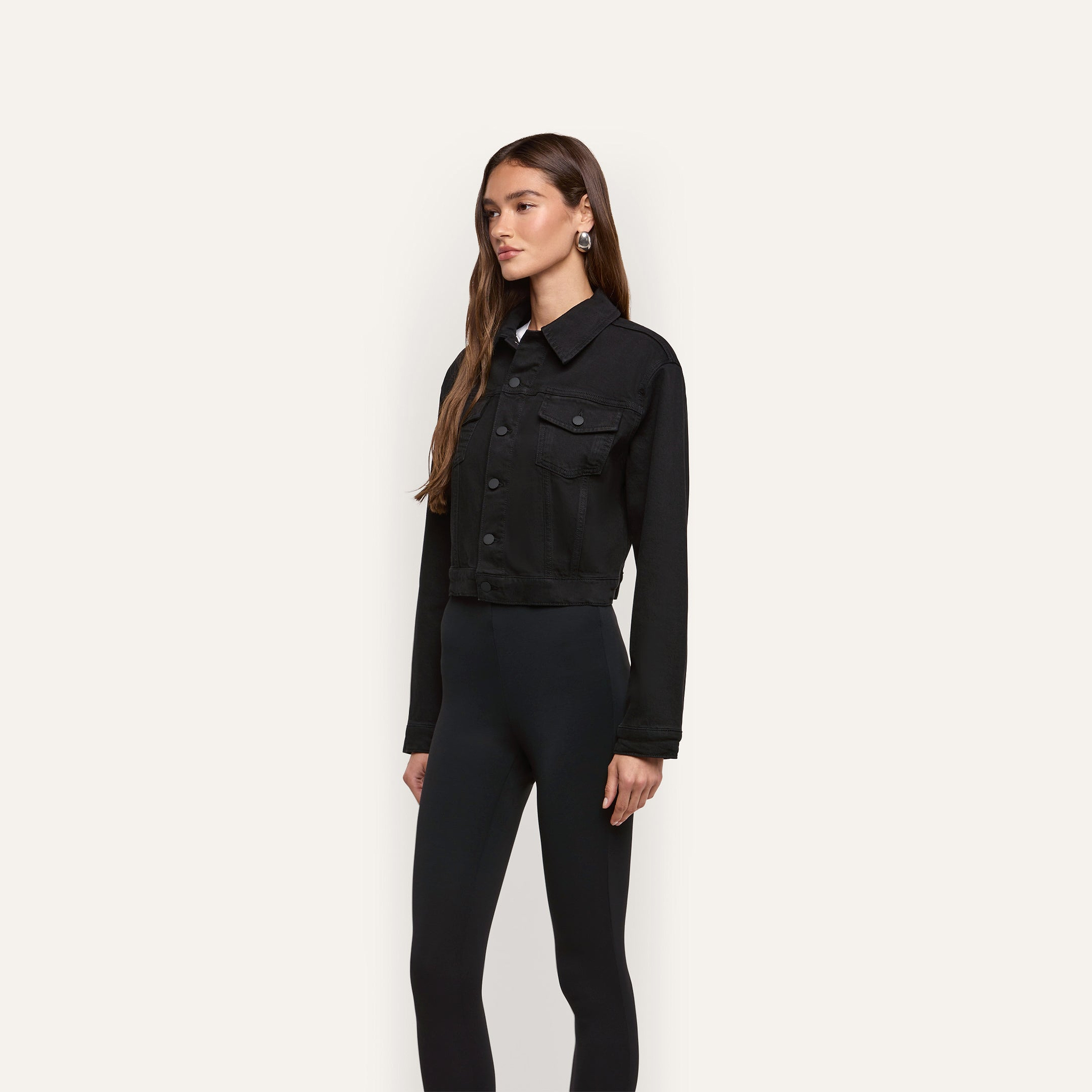 A woman wears a black Alessia Jean Jacket, featuring a cropped length, button-front closure, and two chest pockets, over black leggings.