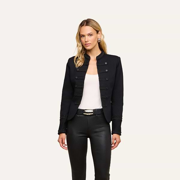 A woman wearing the Victory Jacket, a vintage-inspired black military-style piece, layered over a white top and paired with black pants and a belt, stands against a plain light background.