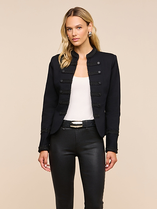 A woman stands facing forward against a neutral beige background, wearing the Victory Jacket—a vintage-inspired black military-style jacket—paired with a white top, black pants, and a black belt featuring a gold buckle.