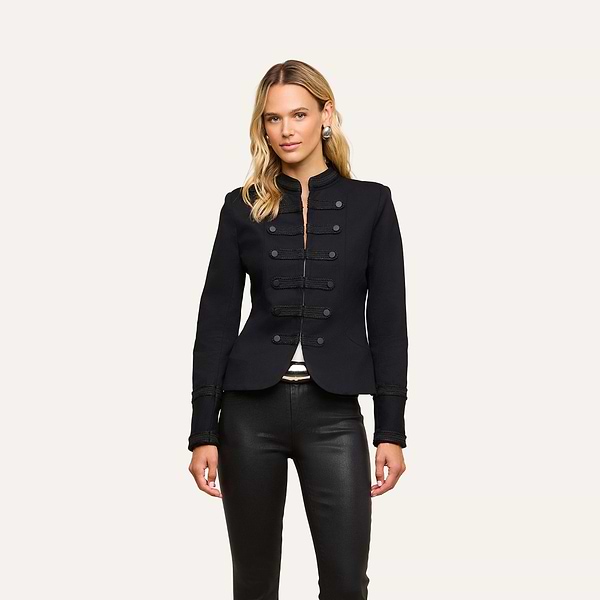 A woman is wearing the Victory Jacket, a fitted black Victorian-style piece with decorative buttons, paired with black leather pants and standing against a plain, light background.