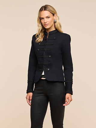 A woman with long blonde hair wears the Victory Jacket— a vintage-inspired black military-style piece— paired with black pants, standing against a plain beige background and looking at the camera with a neutral expression.