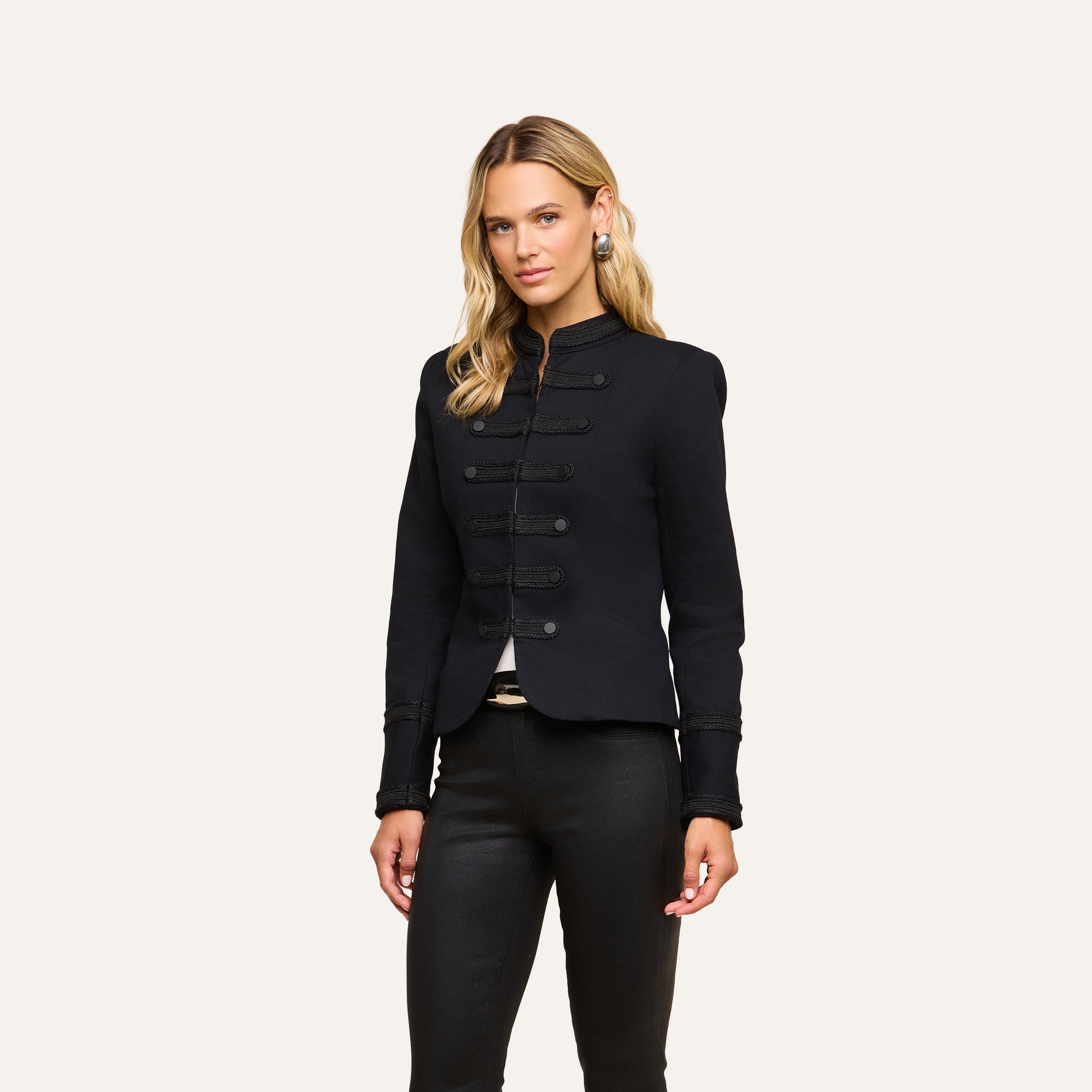 A woman stands facing forward, wearing the vintage-inspired black Victory Jacket with decorative buttons and black pants, against a plain light background.