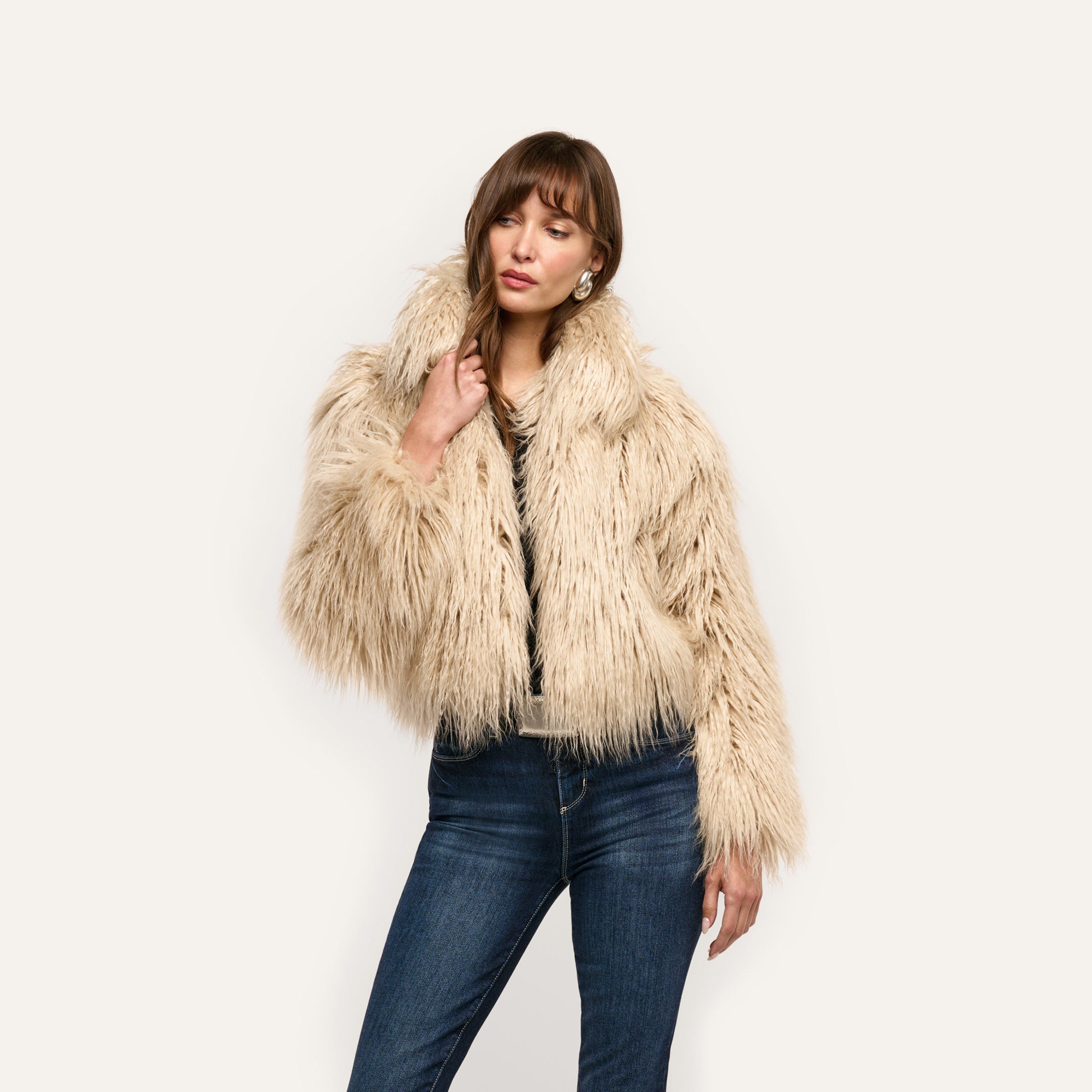 A woman wears a cropped, shaggy Fenella Faux Fur Coat in a neutral tone styled with dark blue jeans.