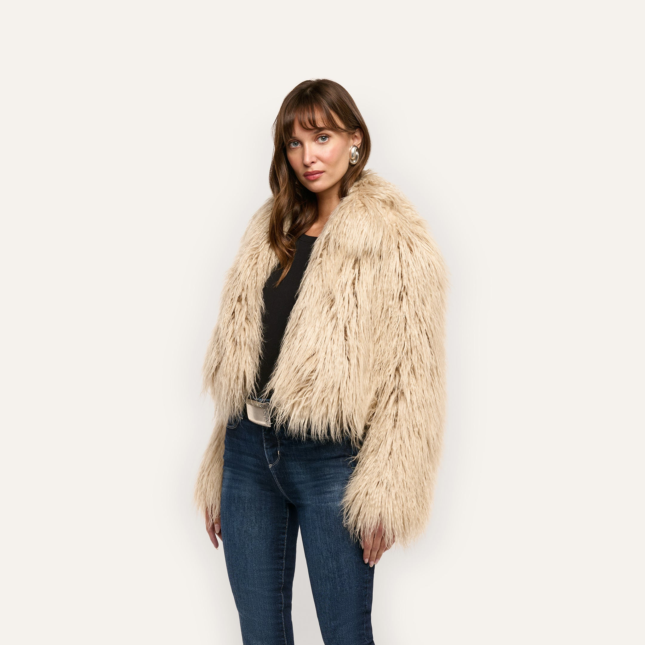 A woman wearing a cropped, shaggy Fenella Faux Fur Coat as statement outerwear over a black shirt and dark blue jeans.
