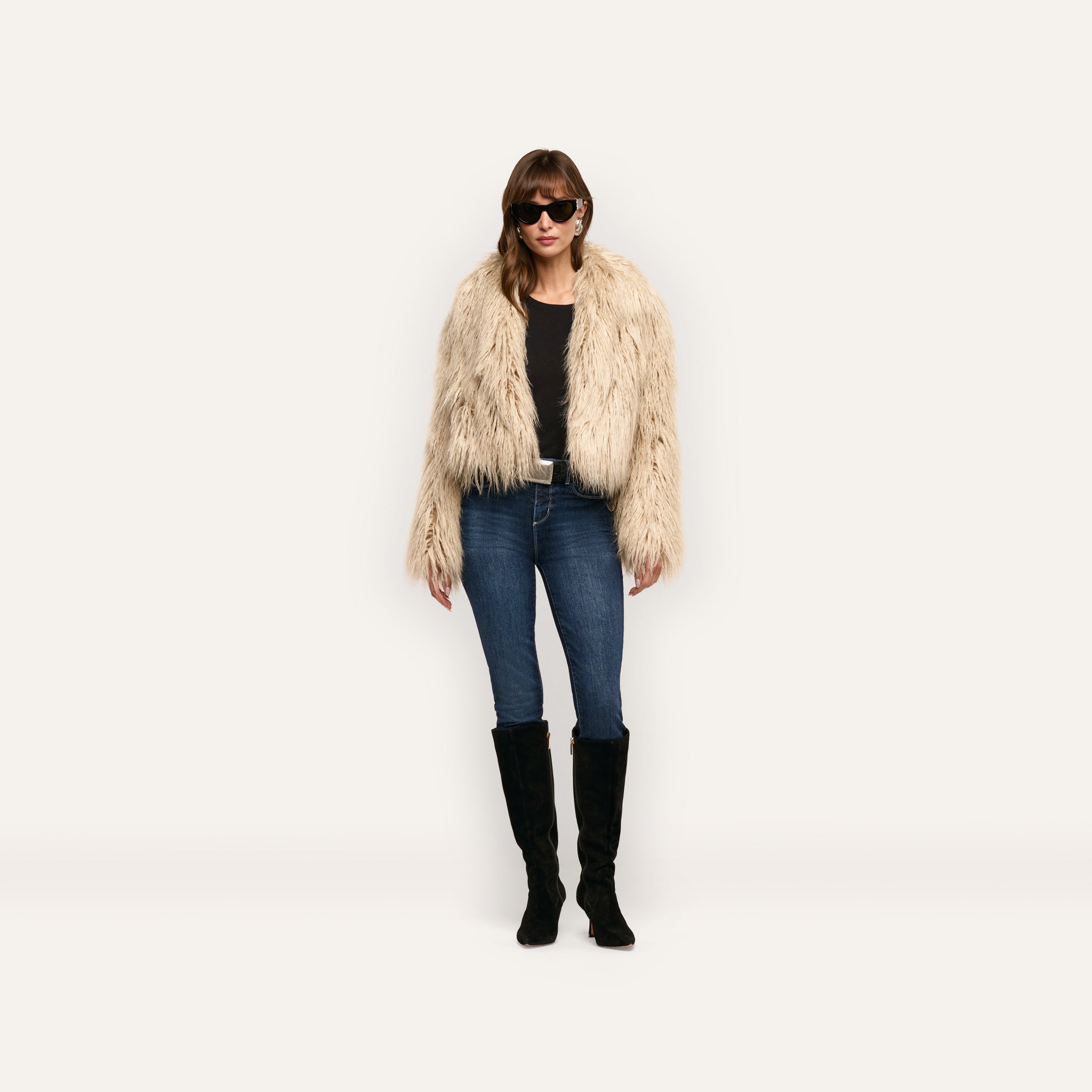 A woman wears a Fenella Faux Fur Coat over a black shirt, blue jeans, and black knee-high boots.