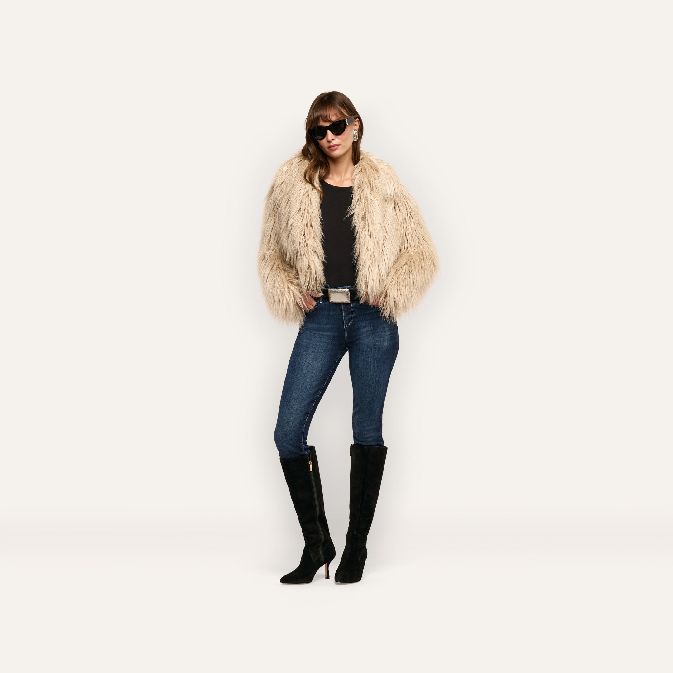 A person wears a cropped, shaggy beige Fenella Faux Fur Coat over a black top, blue jeans, and black knee-high boots, accessorized with sunglasses.