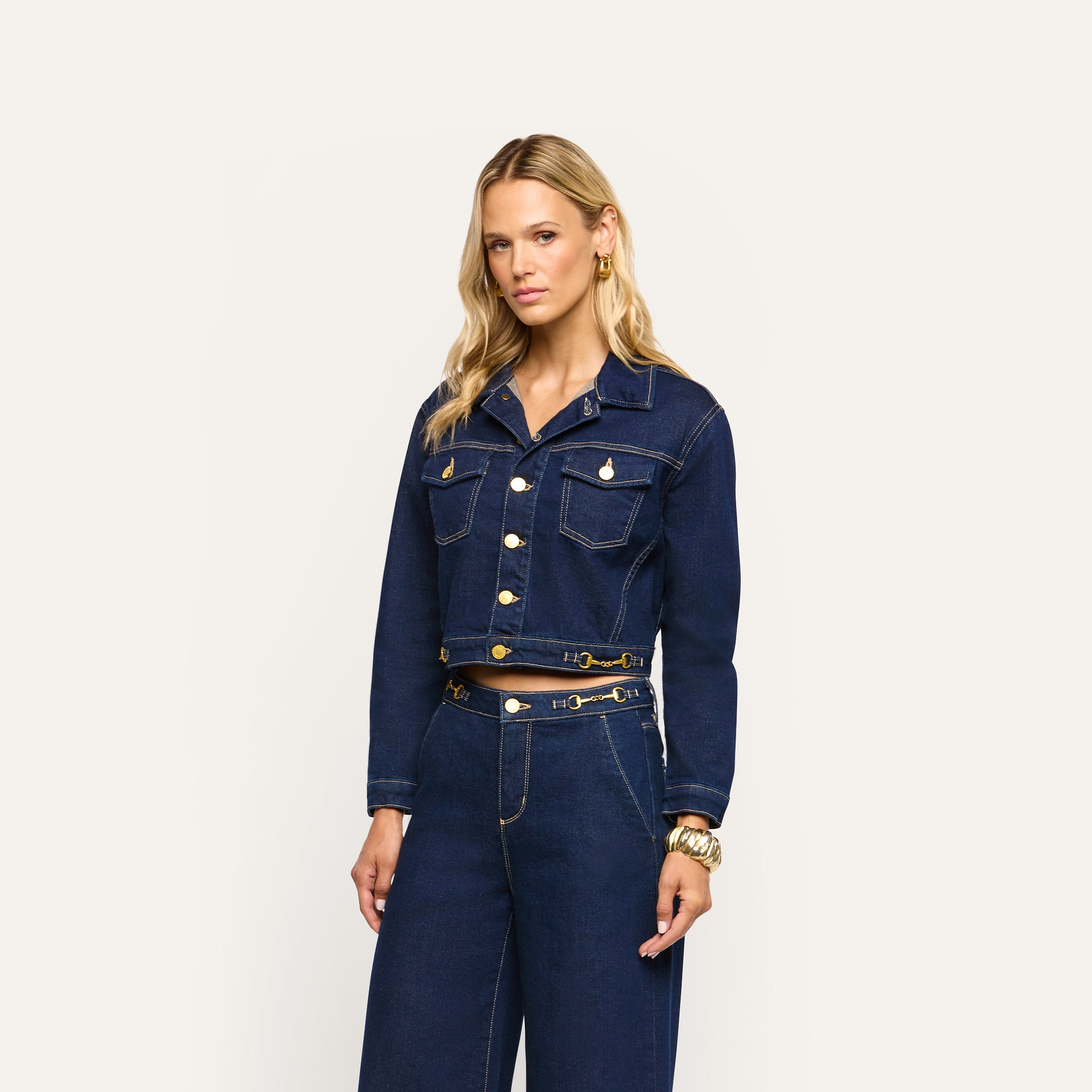 A woman wears an Edin Jean Jacket in dark blue indigo wash with gold buttons, paired with matching high-waisted wide-leg jeans featuring a gold chain detail at the waist.