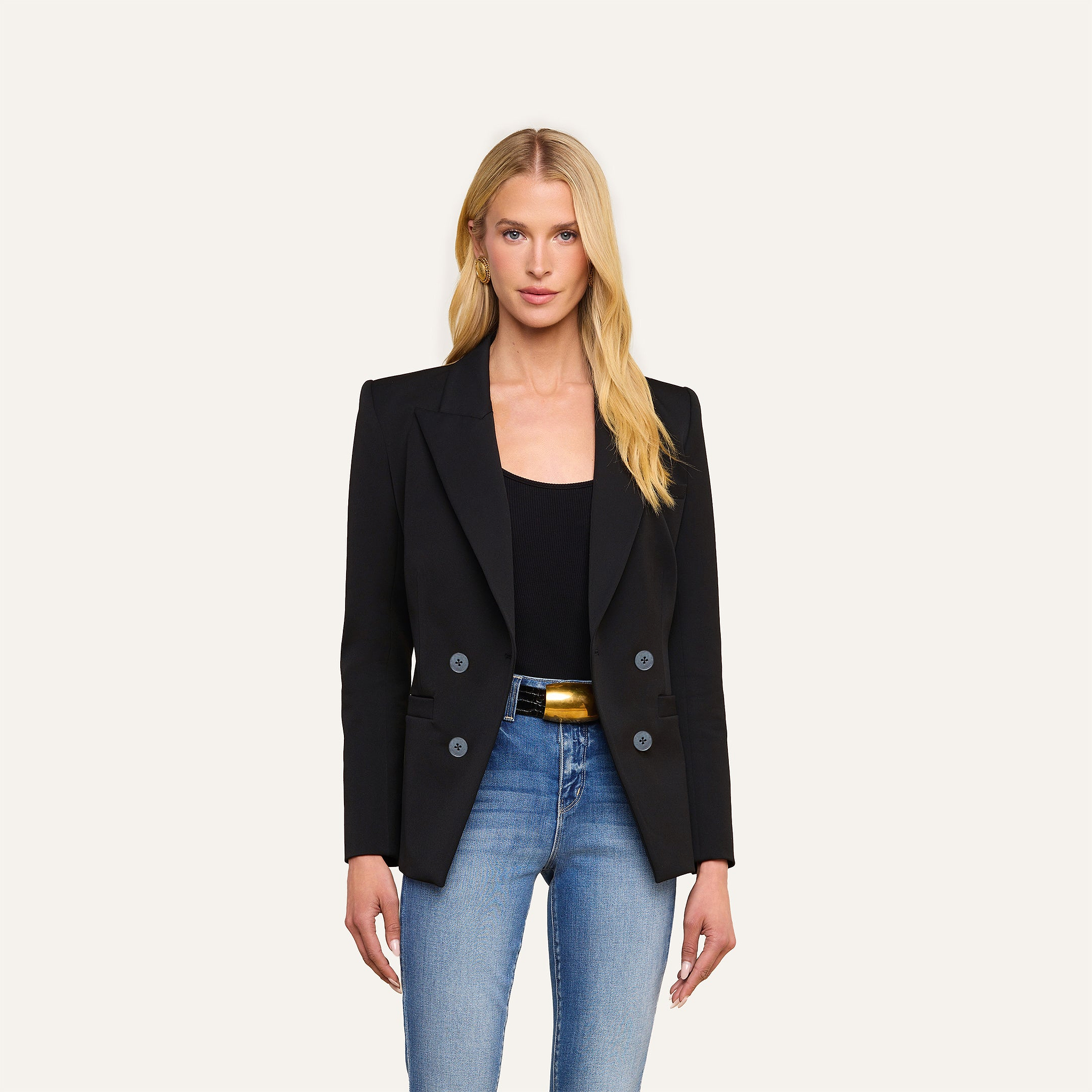 A woman with long blonde hair wears the Kiora Blazer—a black double-breasted jacket with a tailored hourglass shape—paired with a black top, light blue jeans, and a gold-buckled belt, standing against a plain light background.