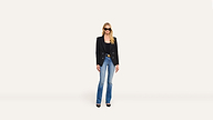 A woman wearing a black blazer, black top, blue jeans, and black heels stands against a plain white background, facing forward and wearing sunglasses.