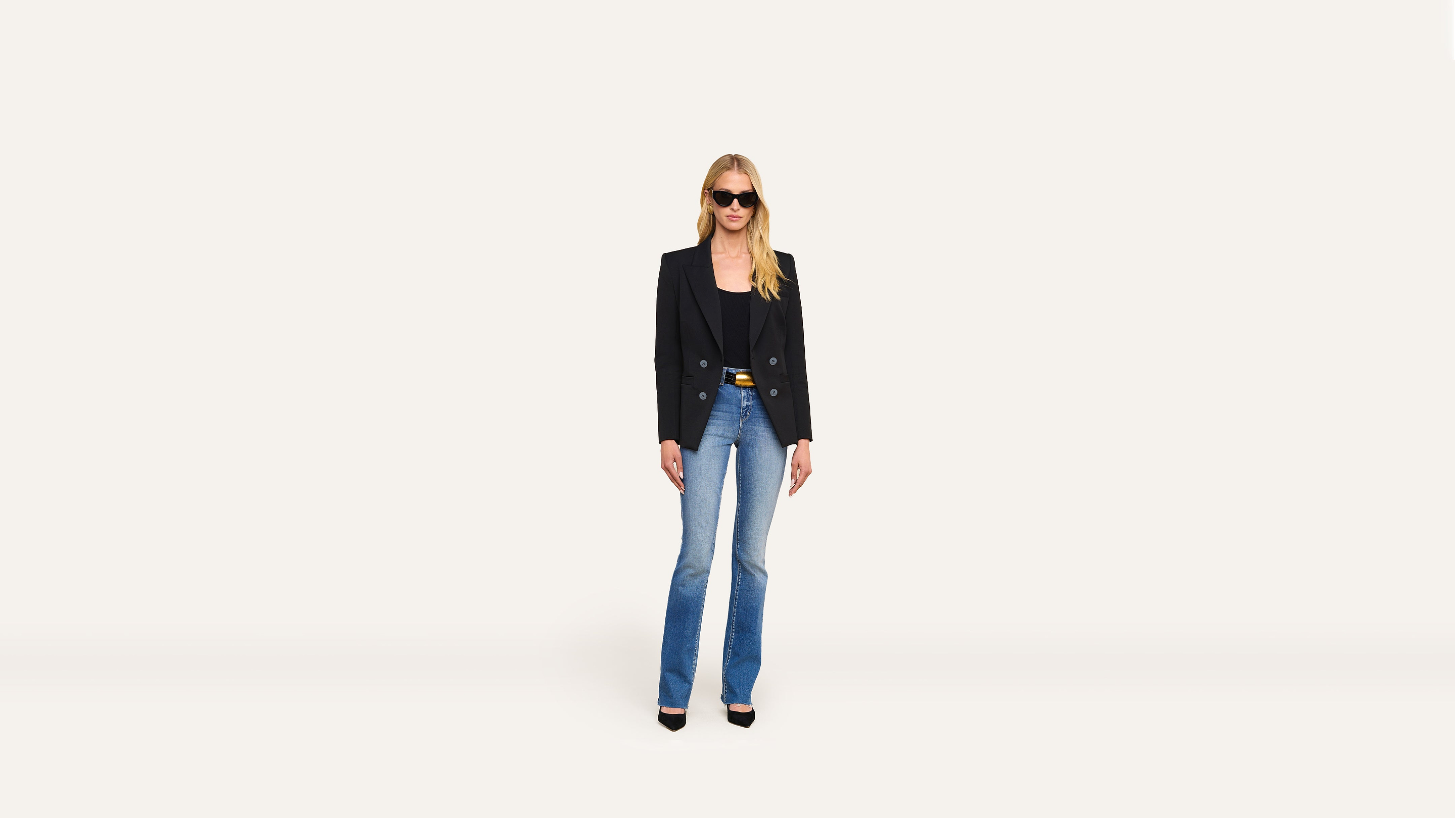 A woman wearing a black blazer, black top, blue jeans, and black heels stands against a plain white background, facing forward and wearing sunglasses.