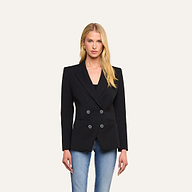 A woman with long blonde hair wears the Kiora Blazer, a black double-breasted jacket with a tailored hourglass silhouette, styled over a black top and blue jeans, standing against a plain light background.