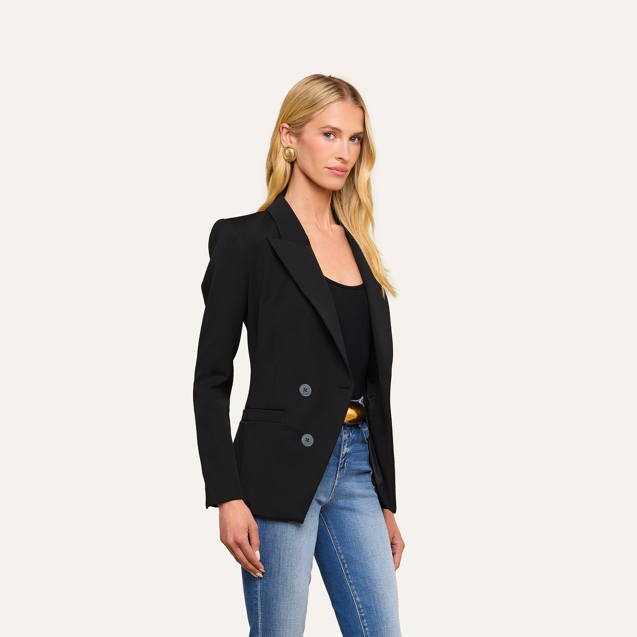 A woman stands against a plain background, wearing the Kiora Blazer over a black top and blue jeans. She has long blonde hair and is facing slightly to the side with a neutral expression.