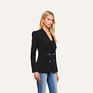 A woman with long blonde hair wears the Kiora Blazer—a black double-breasted style in stretch twill suiting—paired with blue jeans and gold hoop earrings, standing against a plain light background.