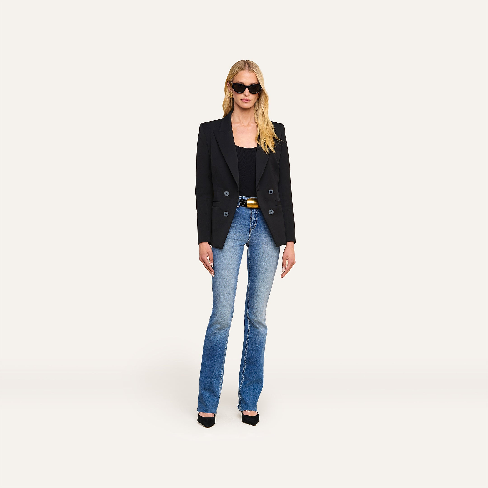A woman stands against a plain white background, facing forward with her hands at her sides, wearing sunglasses, the Kiora Blazer—tailored with an hourglass silhouette—in black, paired with a black top, blue jeans, and black heels.