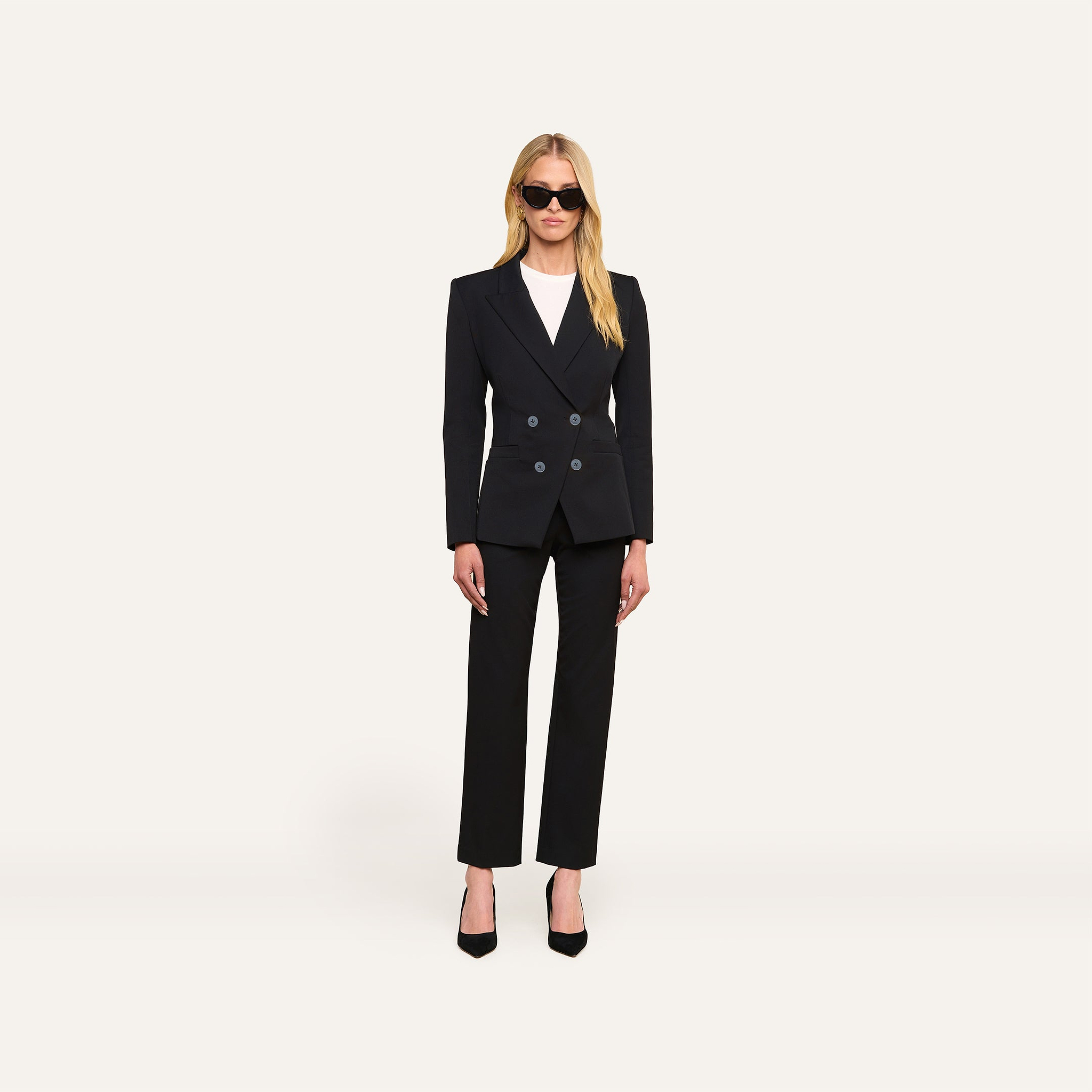 A woman stands facing forward against a plain white background, wearing the Kiora Blazer—a black double-breasted blazer with a tailored hourglass shape—paired with a white top, black heels, and sunglasses.