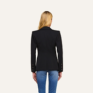 A woman with long blonde hair is shown from the back, wearing the Kiora Blazer—a black fitted piece with a tailored hourglass shape and buttoned sleeves—paired with blue jeans, standing against a plain light background.