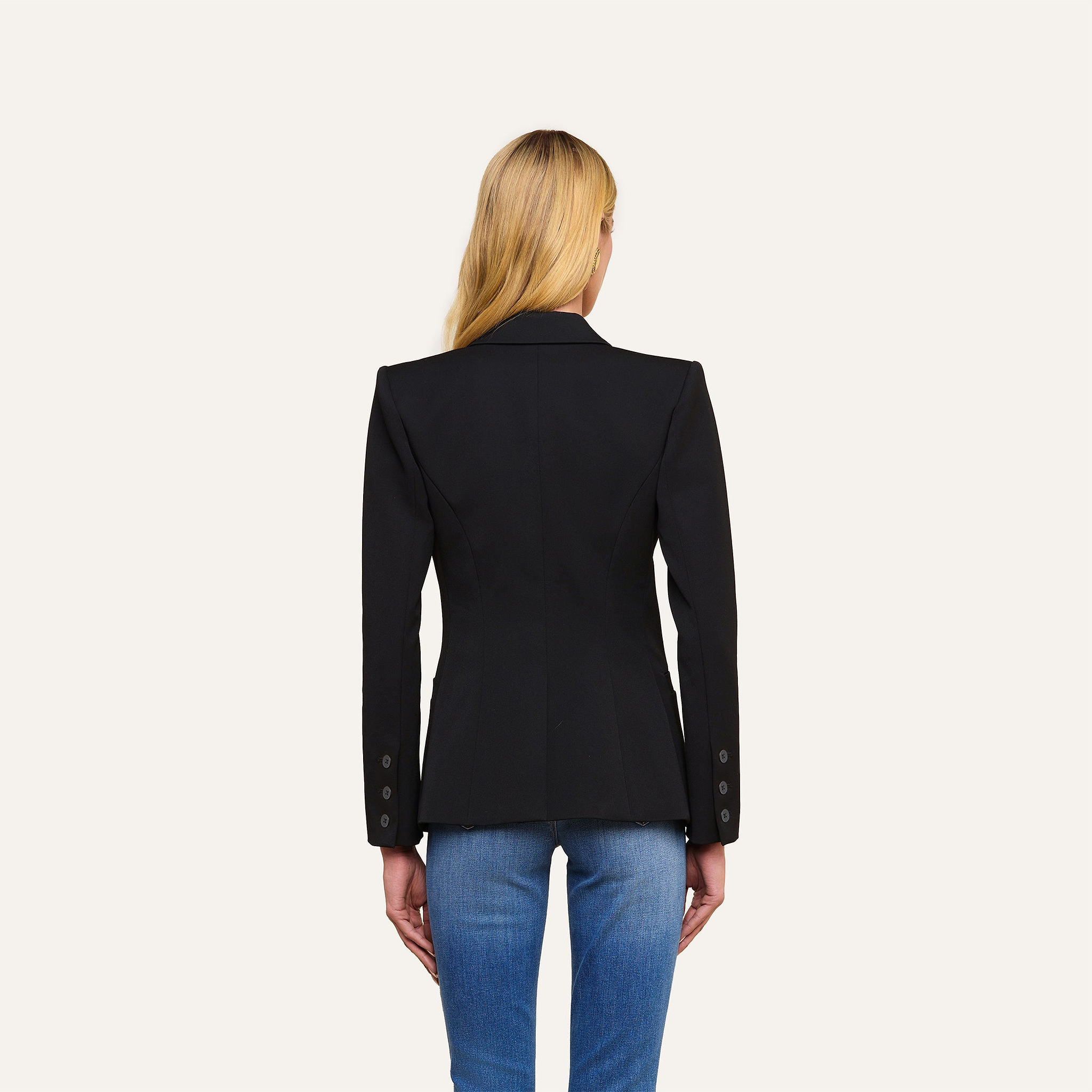 A woman with long blonde hair is shown from the back, wearing the Kiora Blazer—a black fitted piece with a tailored hourglass shape and buttoned sleeves—paired with blue jeans, standing against a plain light background.