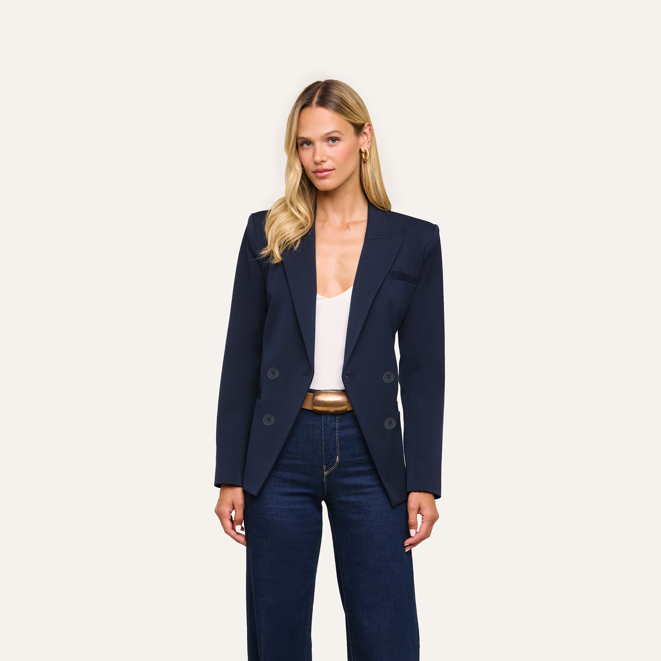 A woman stands facing forward against a plain light background, wearing the Kiora Blazer in navy, styled with a white top and dark blue jeans.