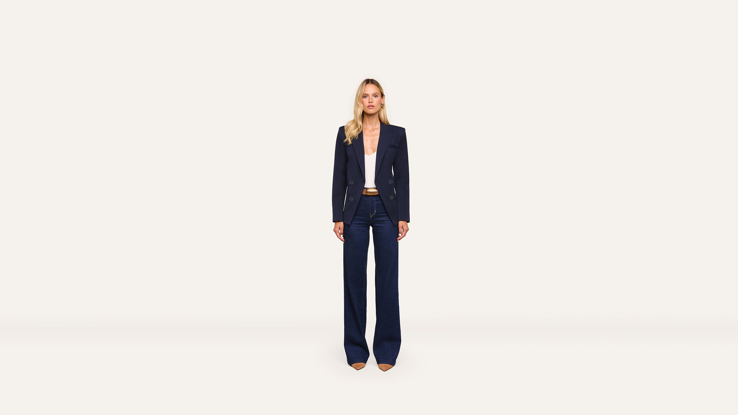 A woman stands facing forward, wearing a navy blazer, wide-leg navy pants, a white top, and tan heels against a plain white background. The tailored blazer adds a polished touch to her ensemble.