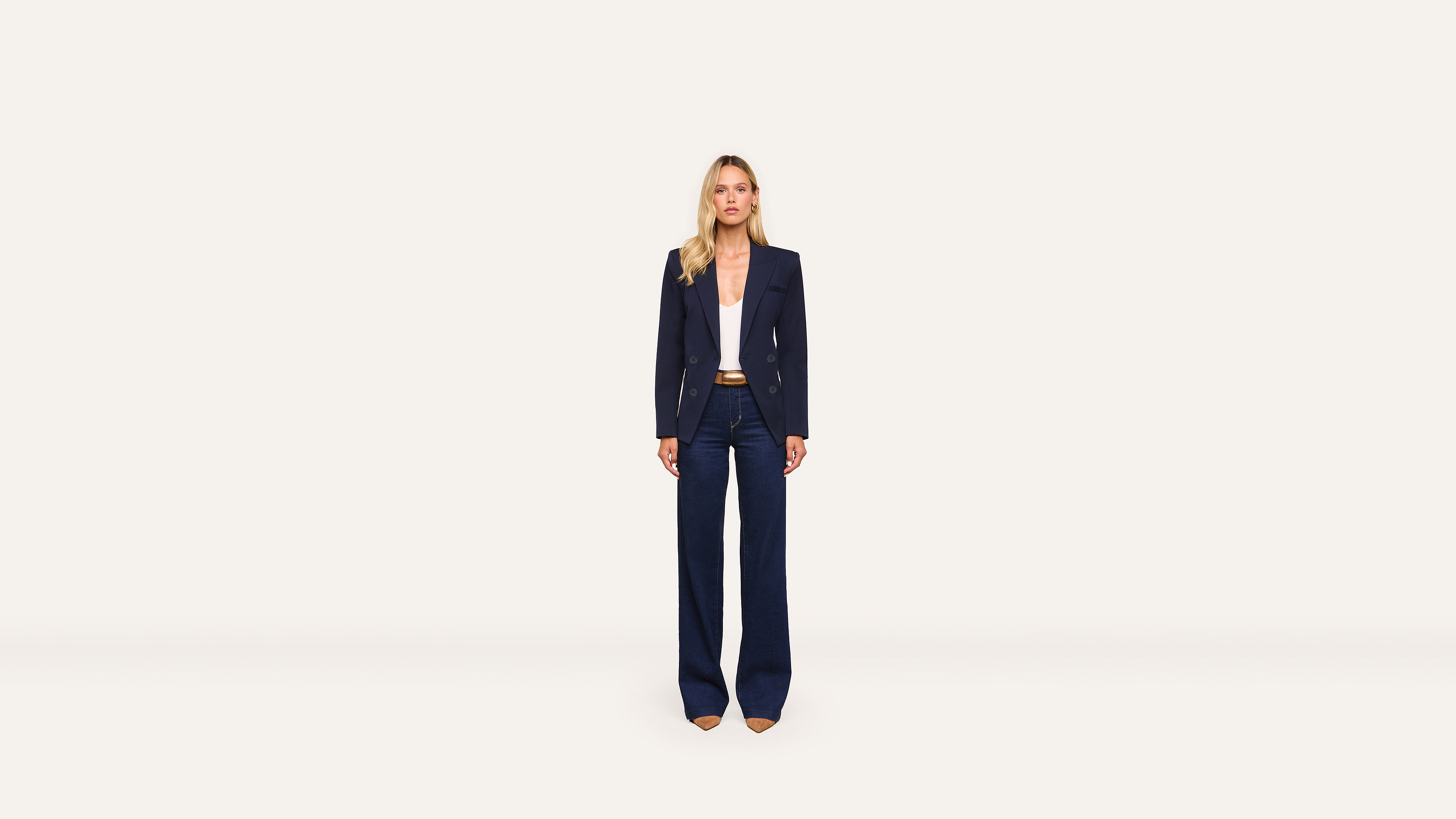 A woman stands facing forward, wearing a navy blazer, wide-leg navy pants, a white top, and tan heels against a plain white background. The tailored blazer adds a polished touch to her ensemble.