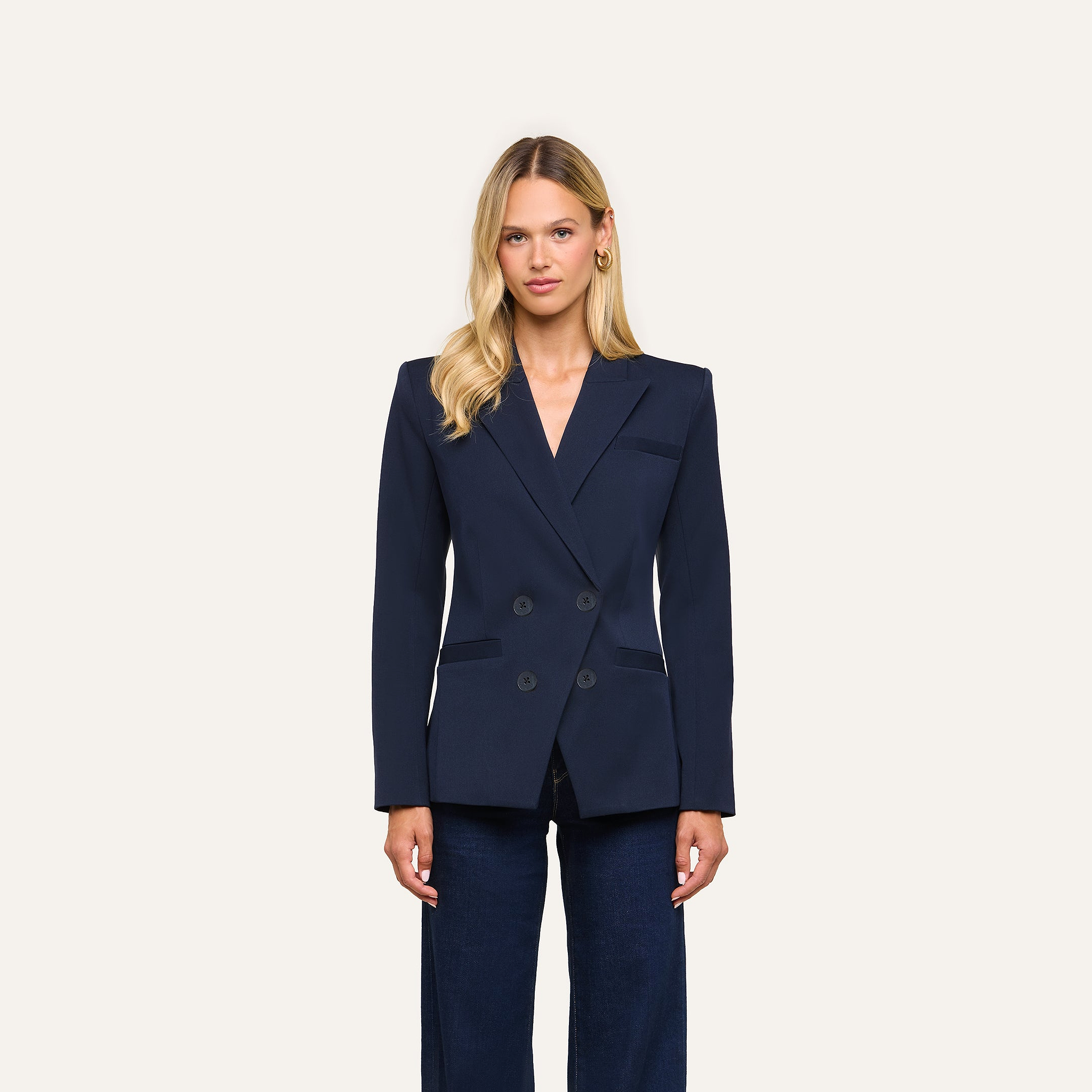 A woman with long blonde hair wears the Kiora Blazer in navy blue and dark pants, standing against a plain light background.