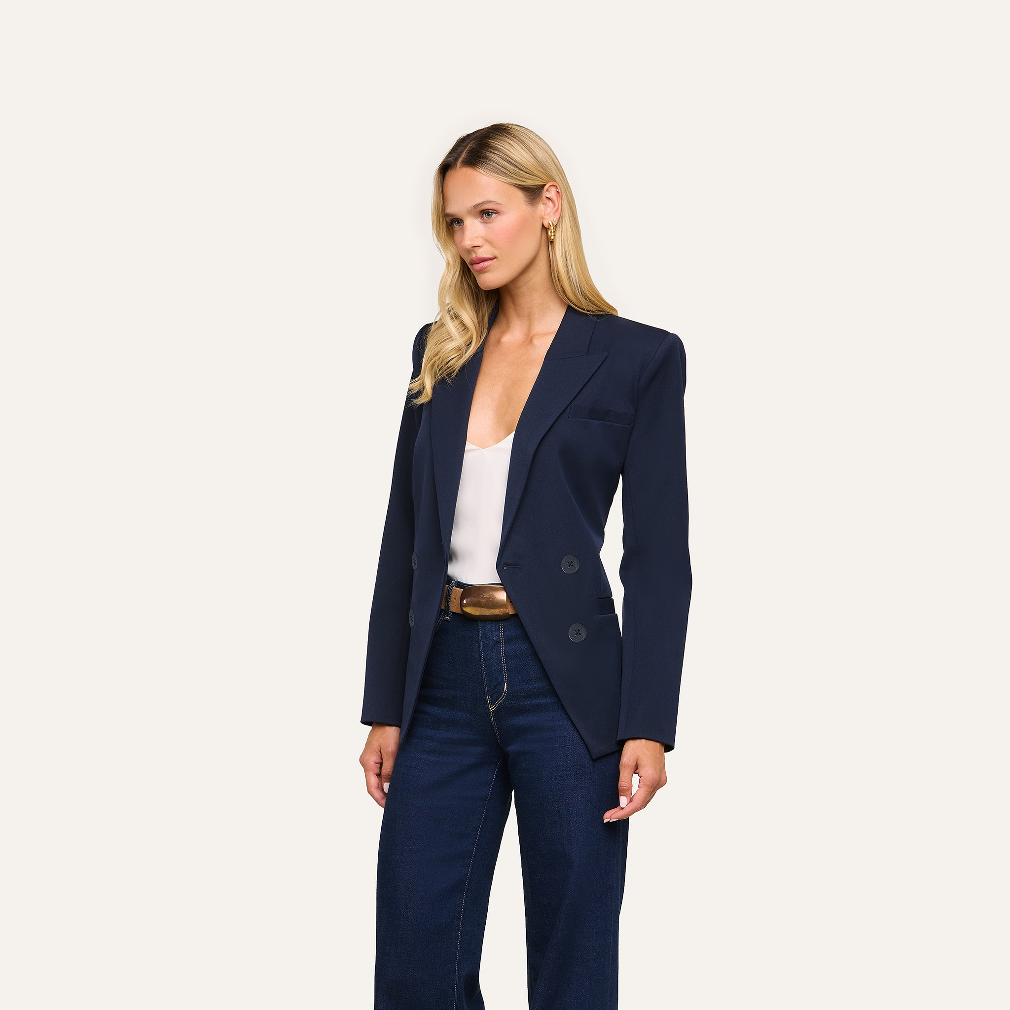 A woman with long blonde hair models the Kiora Blazer in navy over a white top and dark jeans, posing against a plain light background.