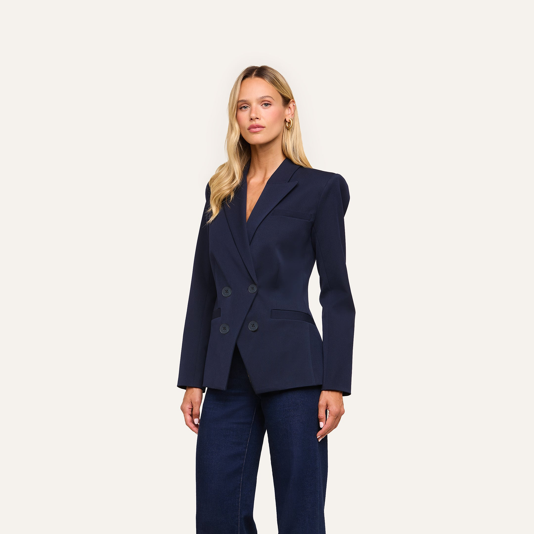 A woman with long blonde hair, wearing the Kiora Blazer in midnight blue paired with dark jeans, stands against a light background and looks confidently at the camera.