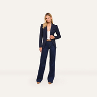 A woman stands facing forward against a plain light background, wearing the tailored hourglass Kiora Blazer in midnight blue over a white top, paired with high-waisted dark blue wide-leg jeans and brown heels.