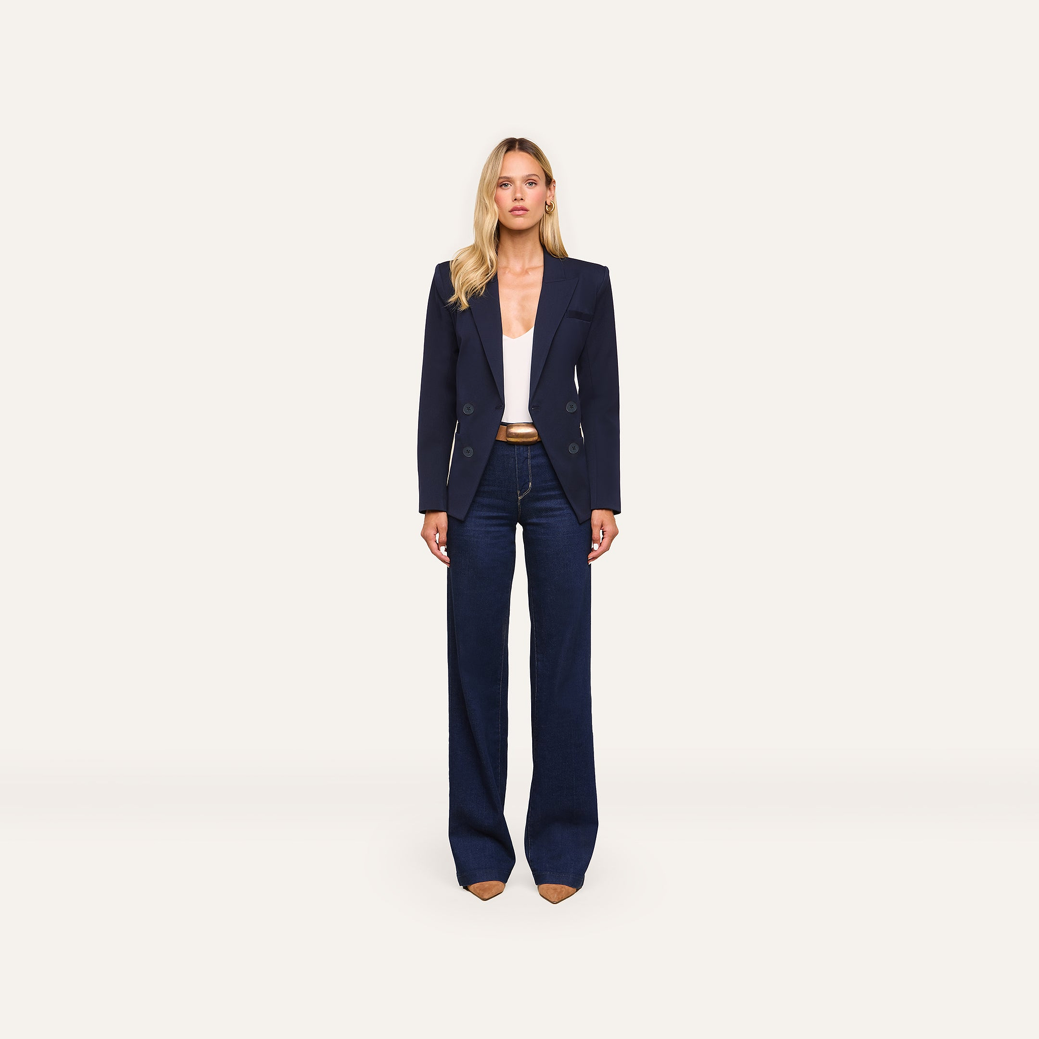A woman stands facing forward against a plain light background, wearing the Kiora Blazer in midnight blue, paired with a white top, dark wide-leg jeans, and beige pointed shoes.