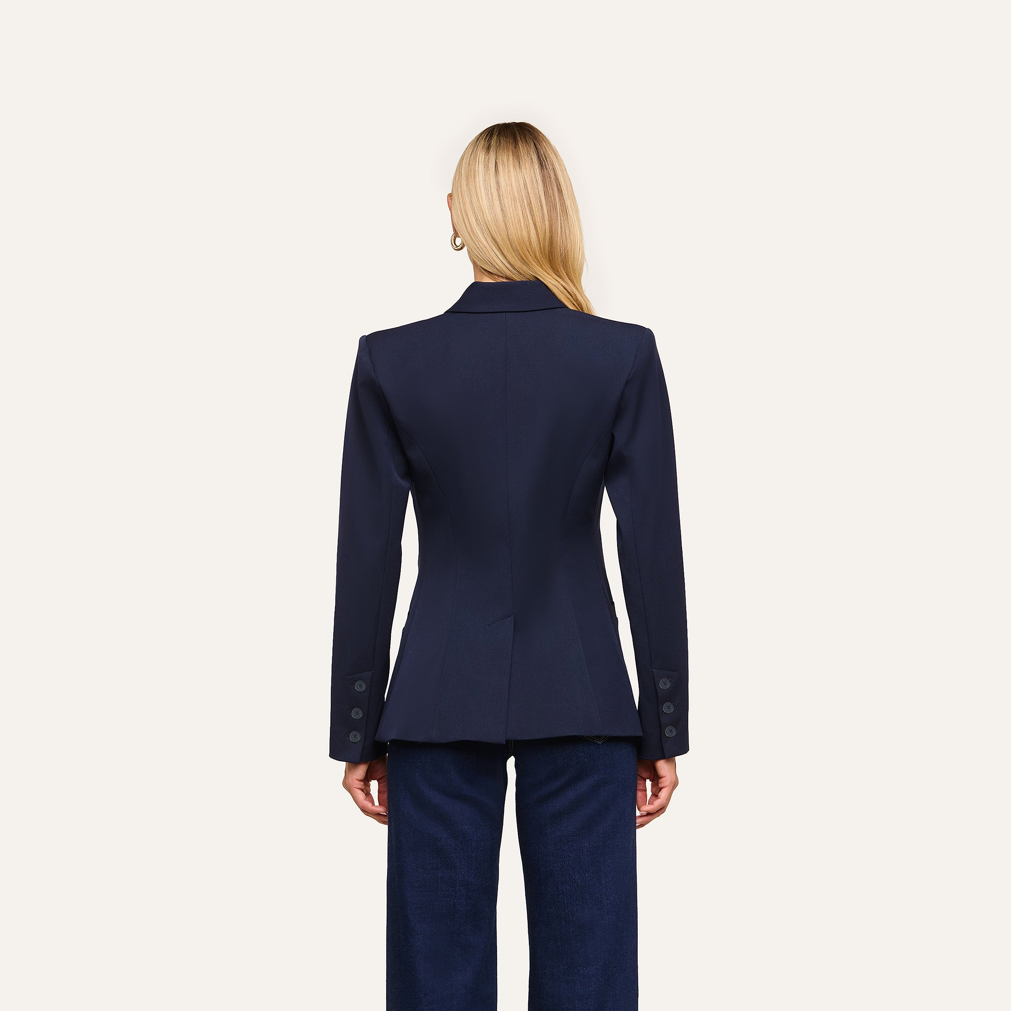 A woman with straight blonde hair, seen from behind, wears the Kiora Blazer and dark trousers against a plain light background.