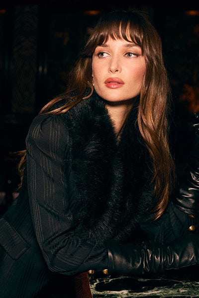 A woman with long brown hair and bangs sits at a marble table, wearing a dark fur-collared coat and black leather gloves, looking thoughtfully to the side in dim, elegant lighting.